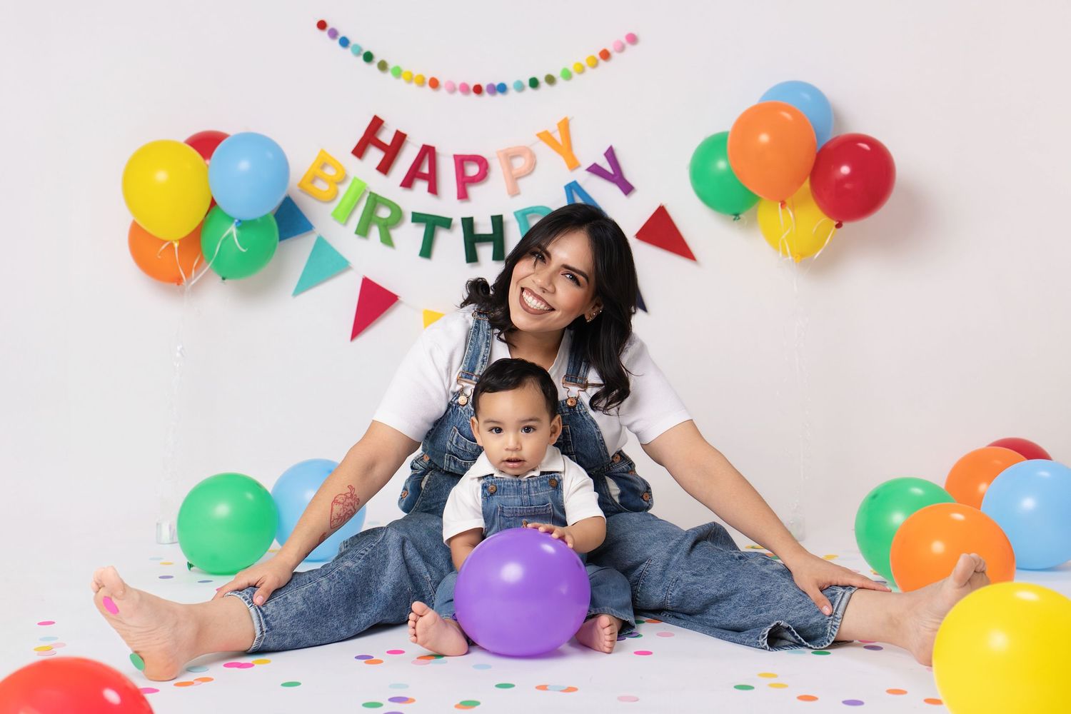 Mother and baby celebrate first birthday with colorful balloons and Happy Birthday banner in a festive photo shoot.