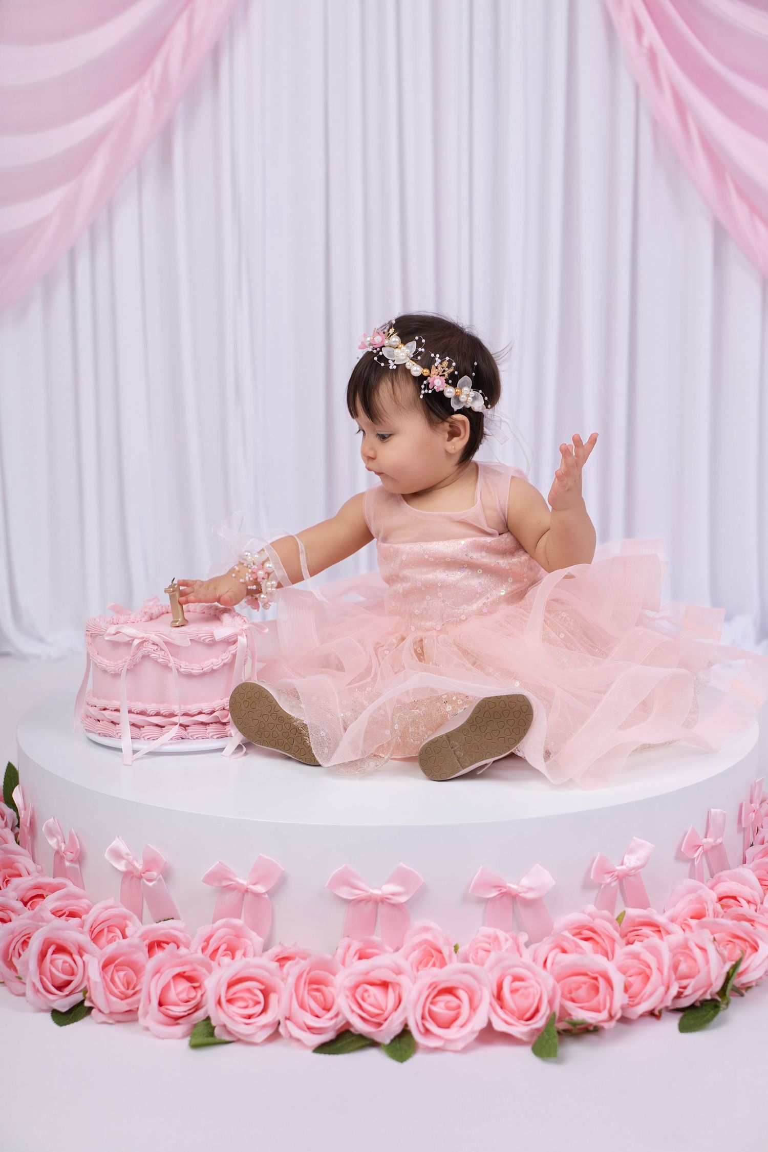 Baby girl in pink tutu dress with floral crown sits on decorated platform touching pink birthday cake surrounded by roses.