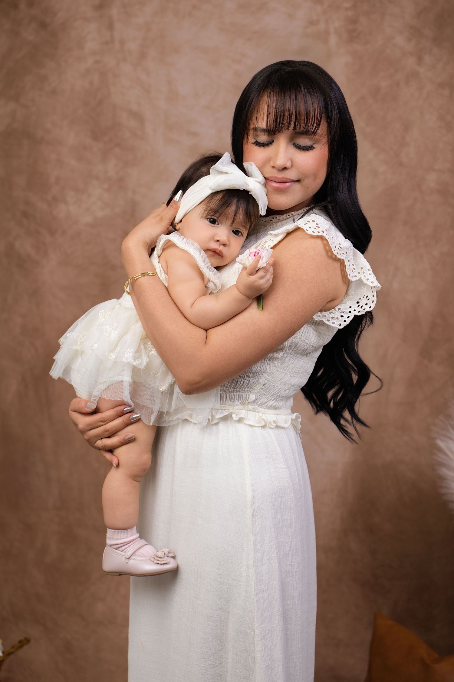 A young woman in a white dress tenderly holds a baby girl in a matching white outfit with a bow headband.