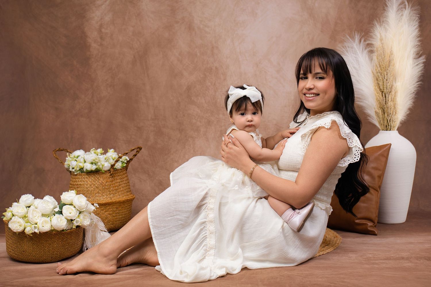 A smiling mother in a white dress holds her baby girl with a bow headband, surrounded by white flowers in a studio setting.