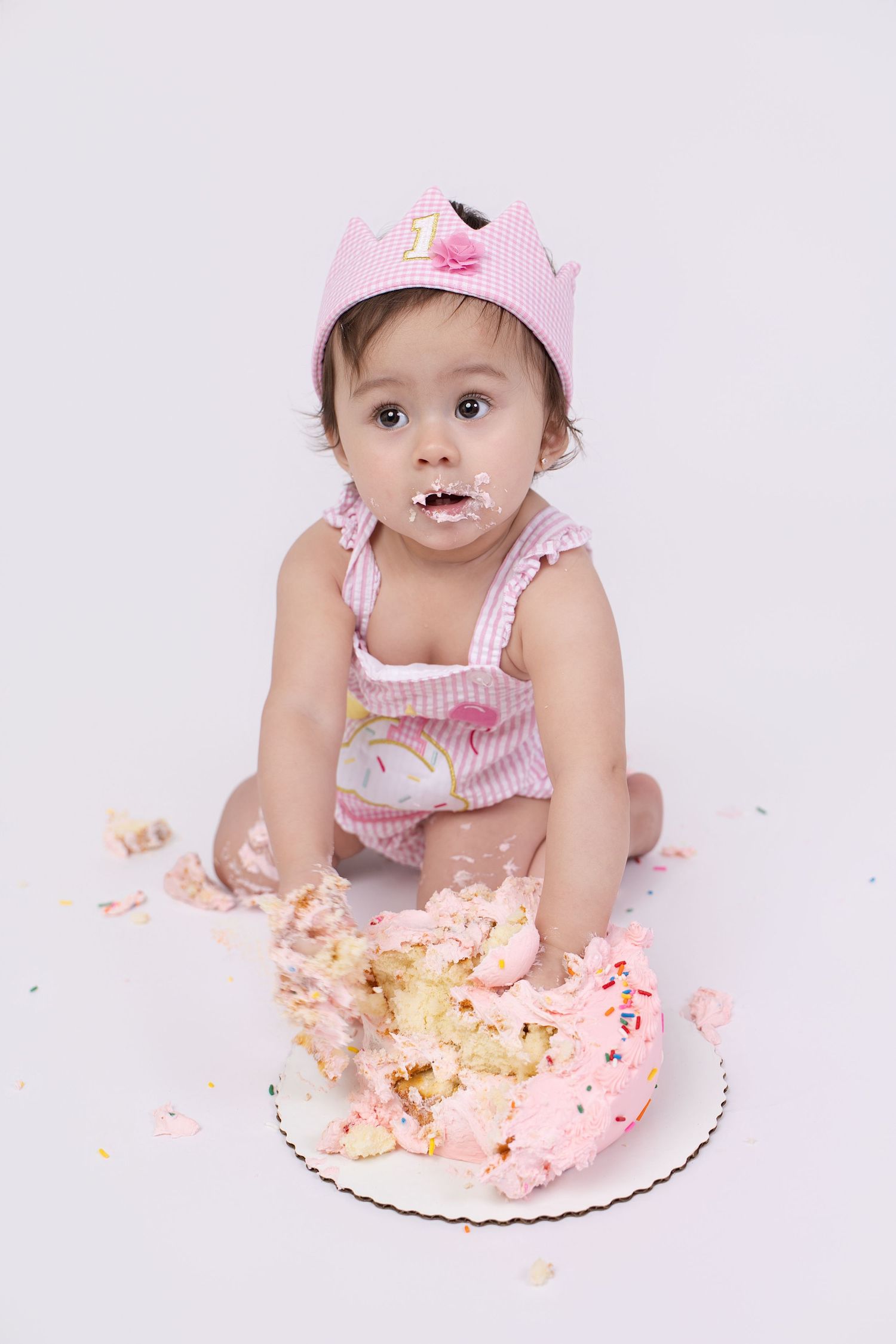 Baby girl in pink crown smashes first birthday cake, frosting on face, during classic cake smash photo session.