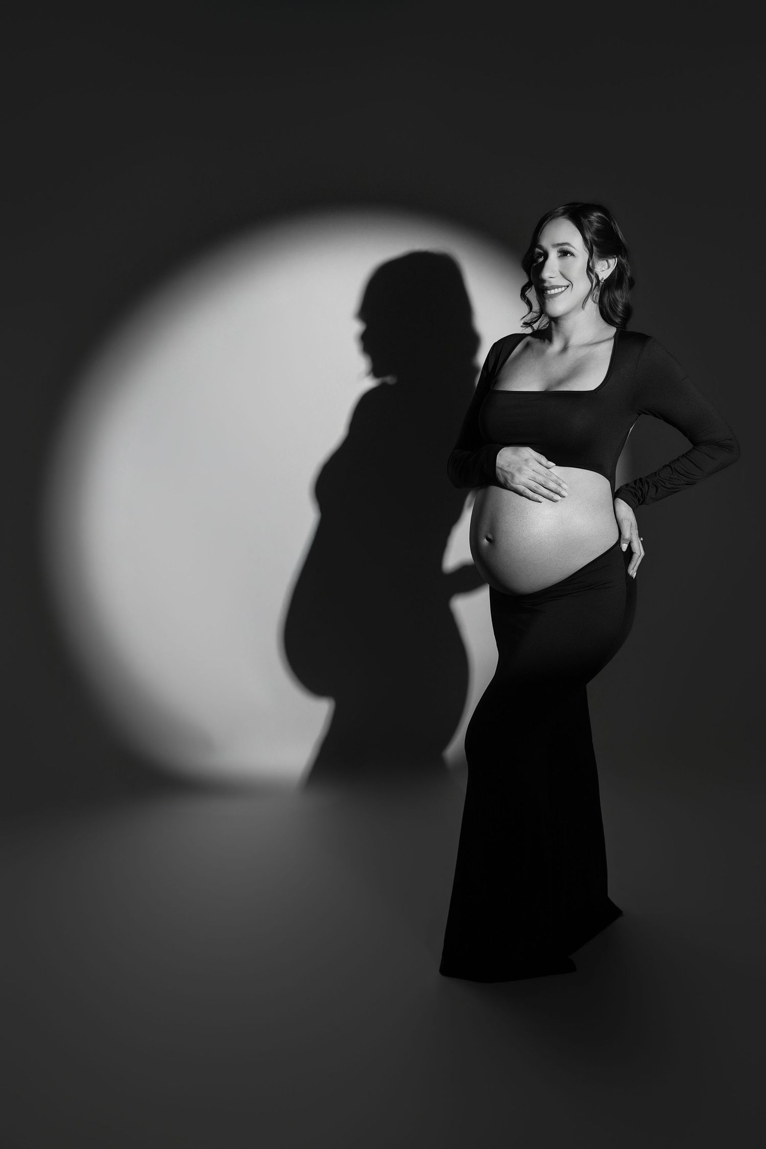 Black and white maternity photo of a smiling pregnant woman in a dark gown, cradling her belly with a dramatic spotlight shadow.