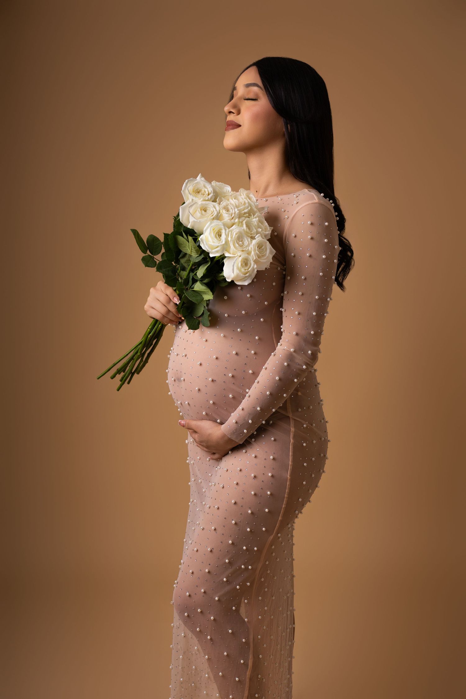 Figure in sheer pink pearl-embellished gown holds white roses against brown backdrop in elegant maternity photoshoot.