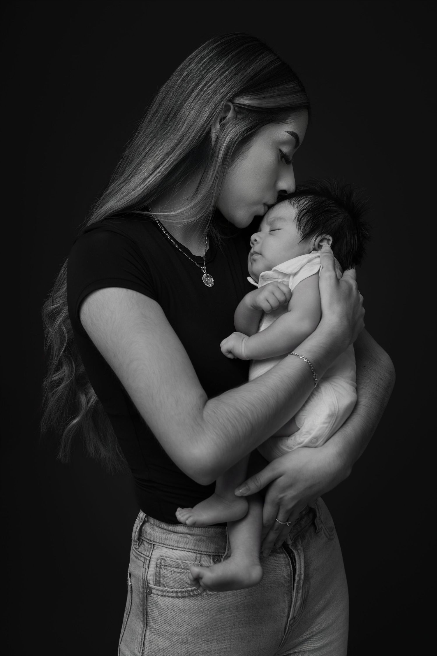 Black and white portrait of a young mother tenderly kissing her sleeping newborn baby while cradling them in her arms.