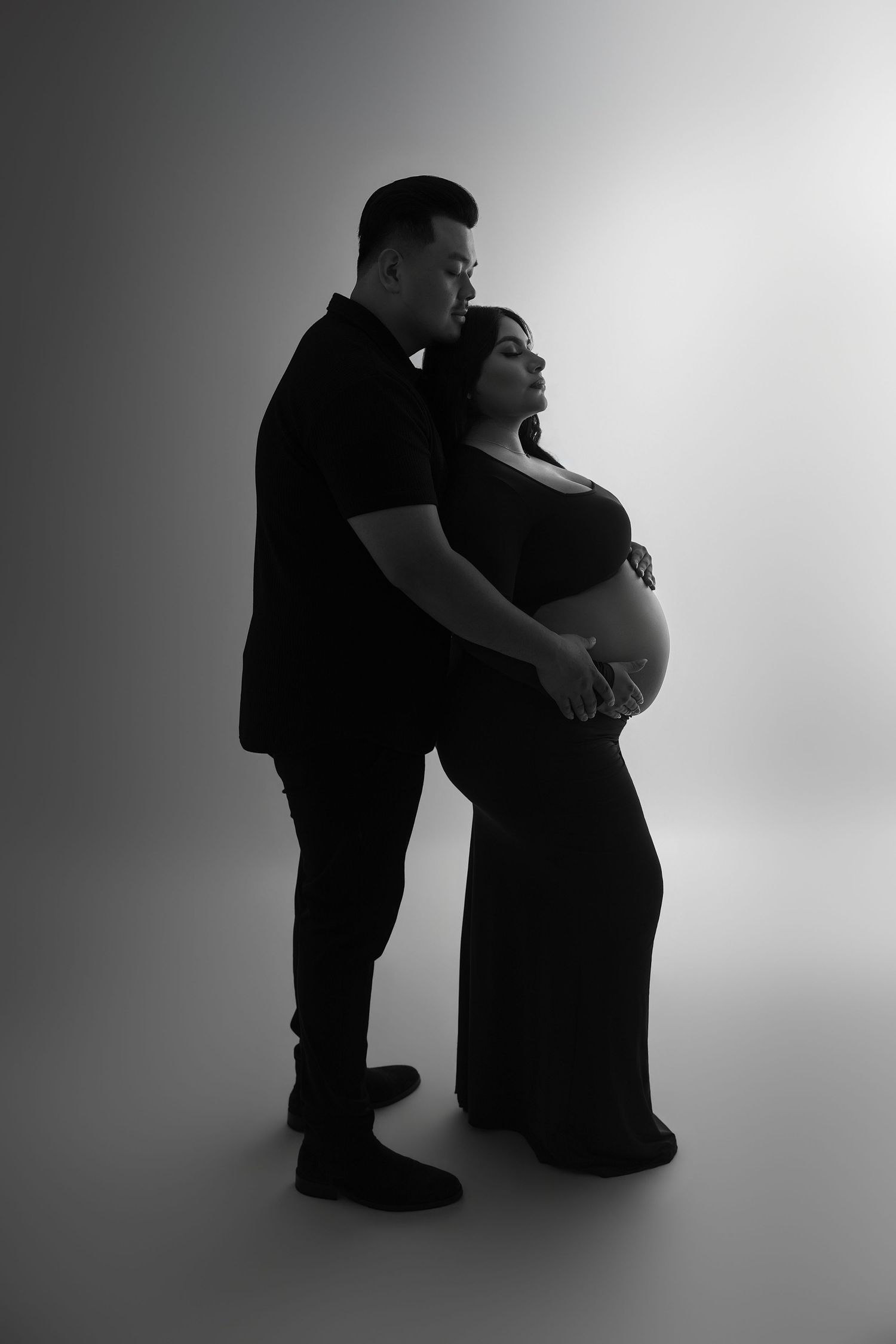 Black and white maternity photo of a couple in dark clothing, man embracing pregnant woman from behind in studio.