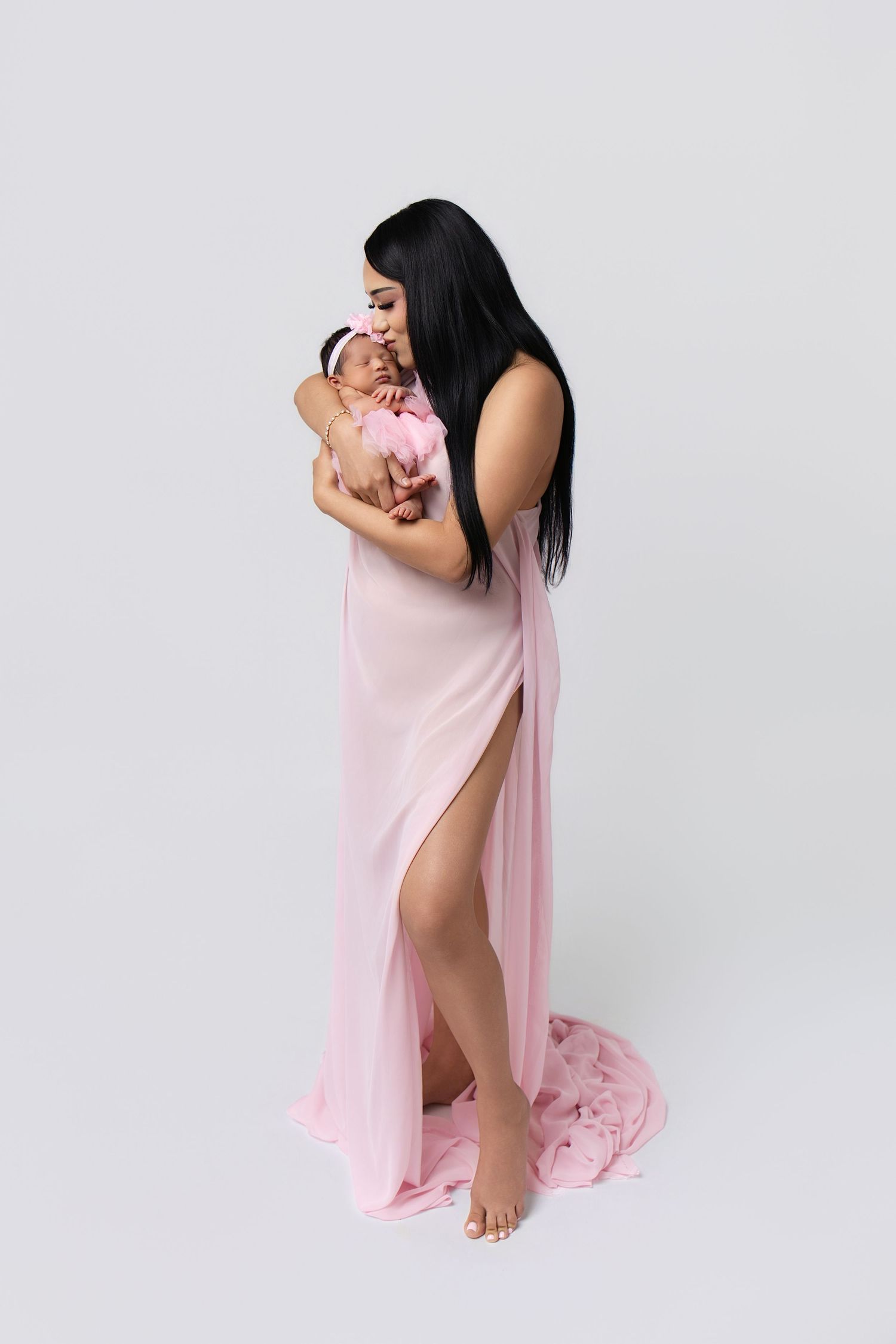 A mother in a pink gown holds her newborn baby, both dressed in pink, against a white background.