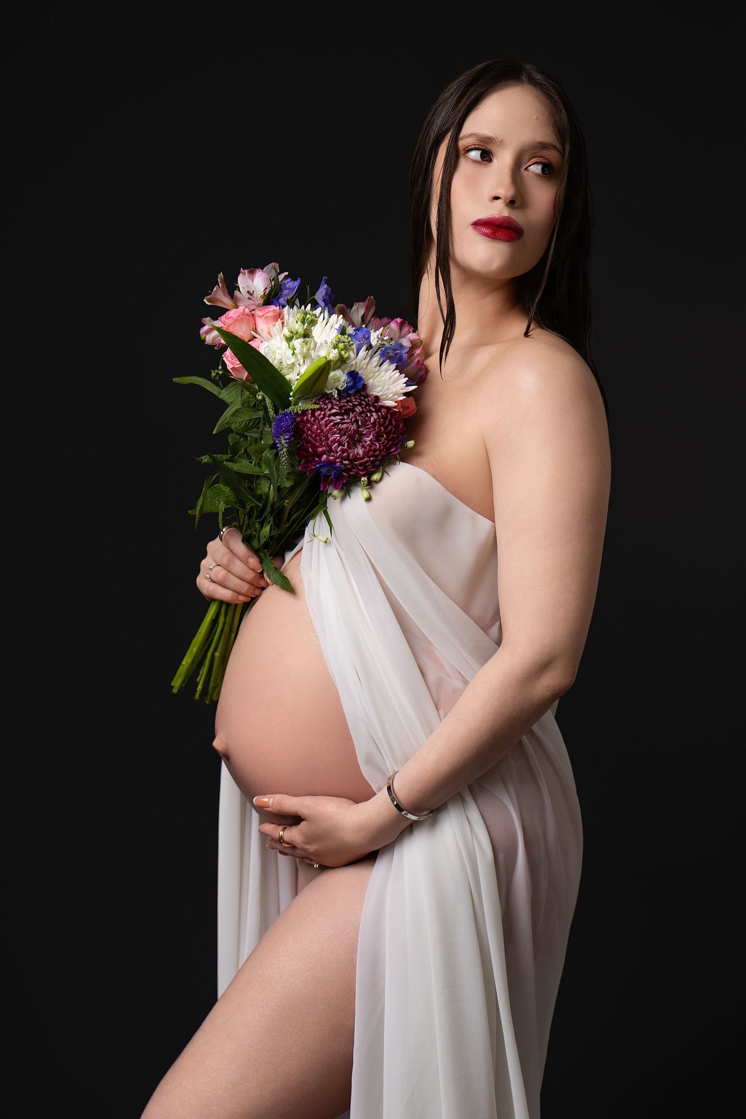 Pregnant woman in white sheer fabric holding colorful flower bouquet, posing against dark background.