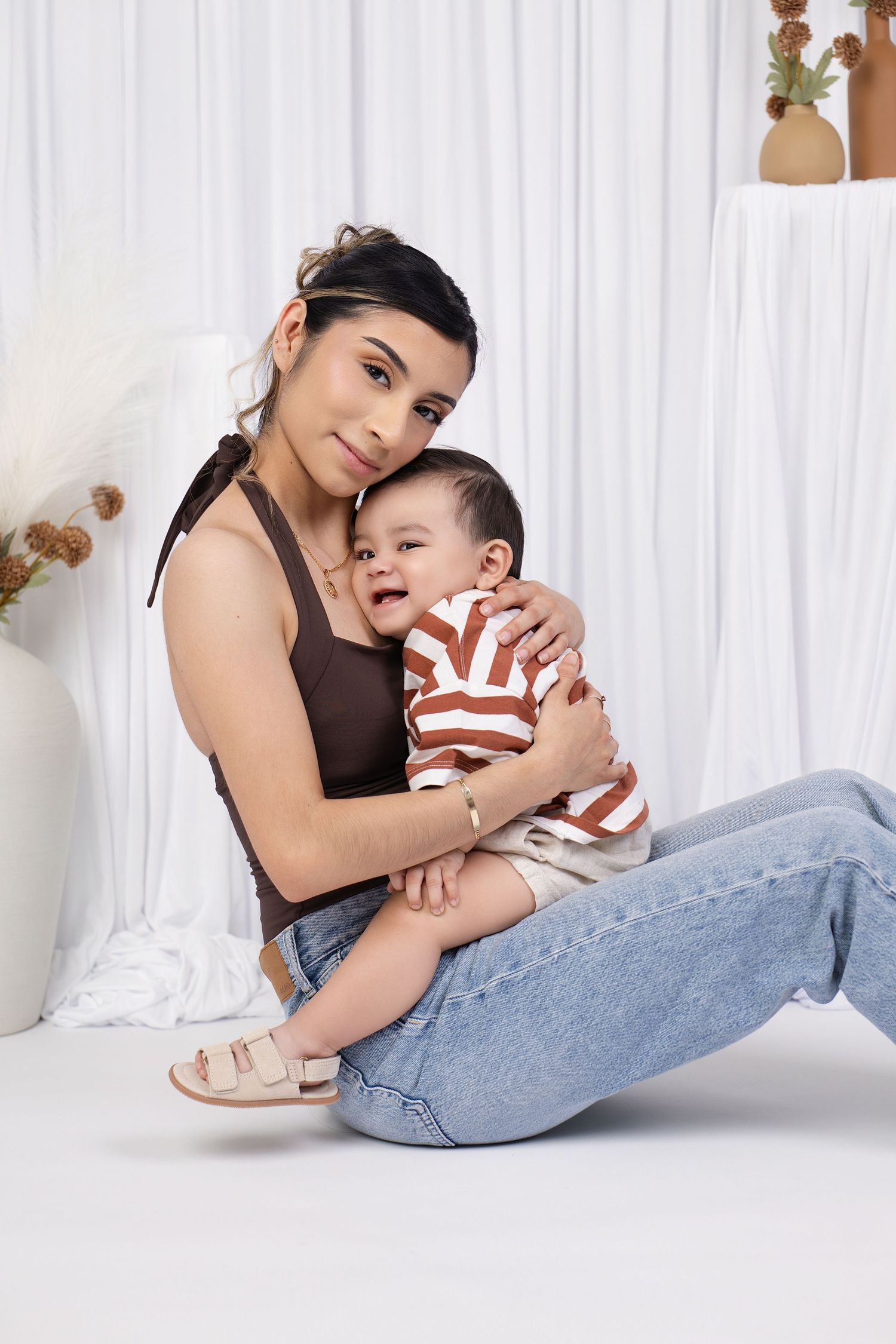 Young mother in brown crop top and jeans sitting on floor holding smiling baby in striped shirt.