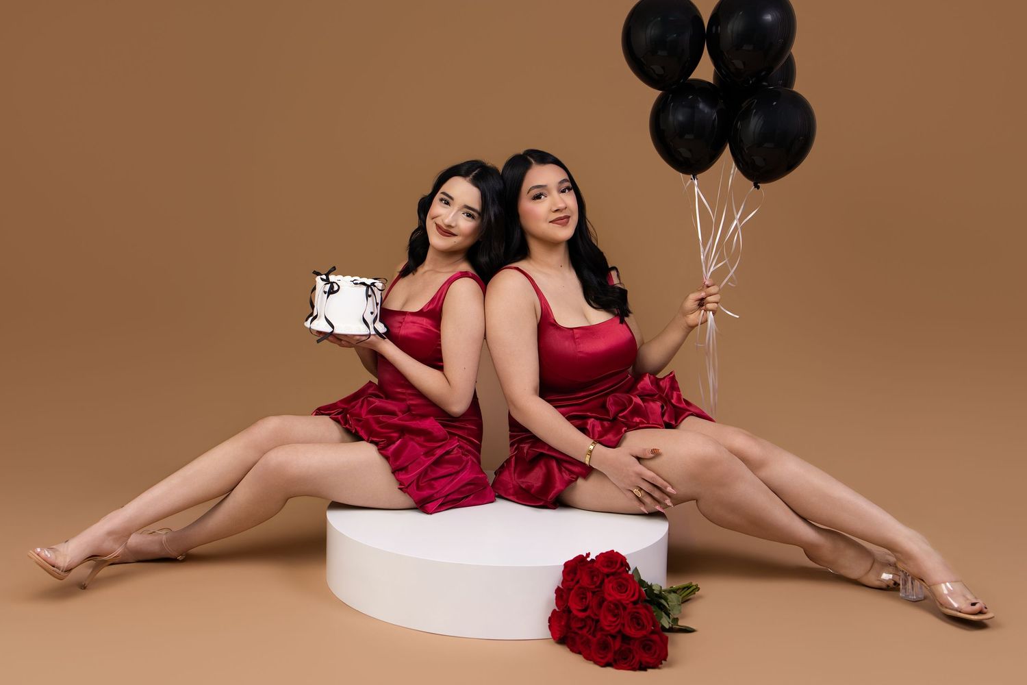 Two women in red dresses sit back-to-back holding a cake and black balloons with red roses on beige background.