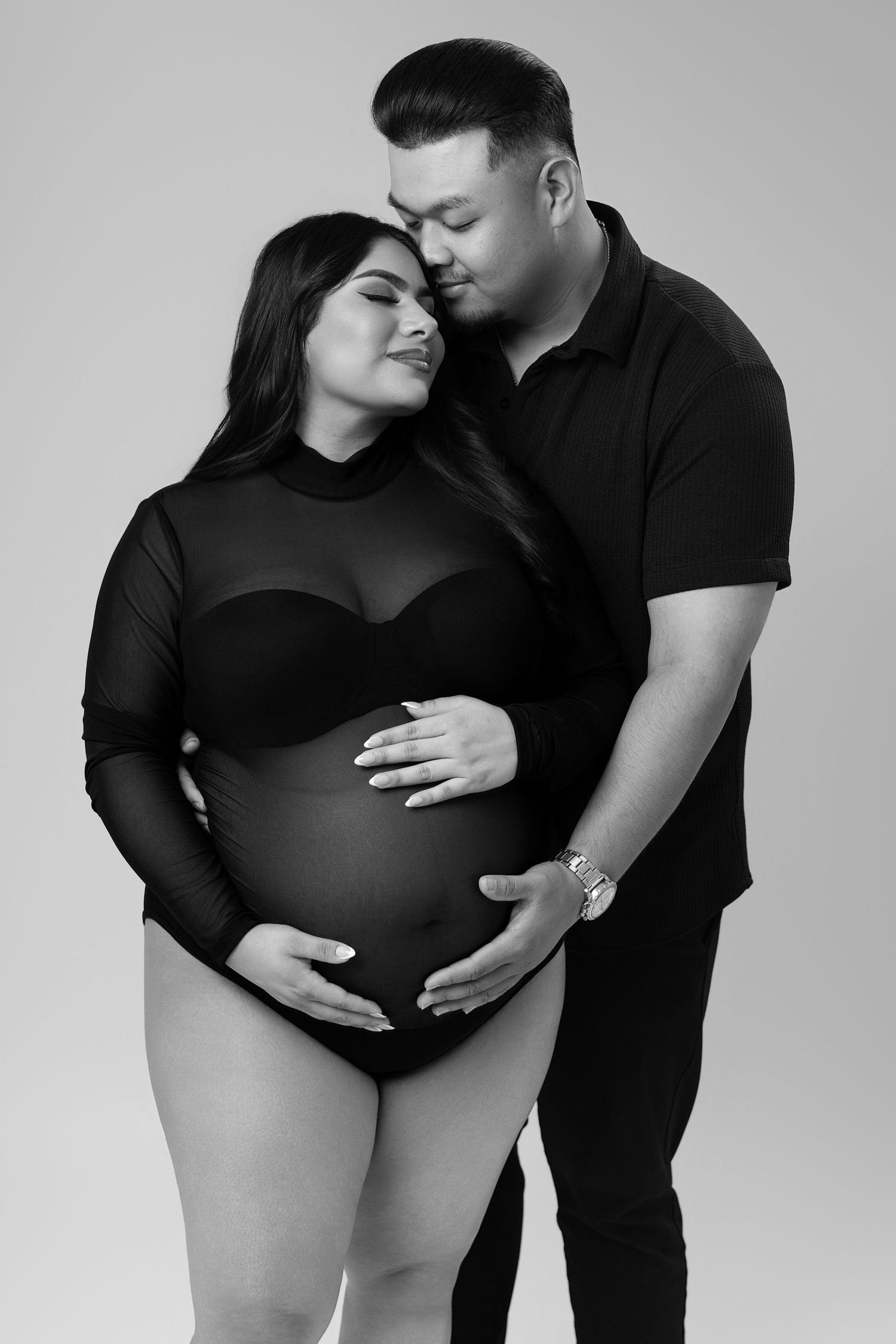 Black and white maternity photo of a couple, man kissing pregnant womans head while both cradle her baby bump.