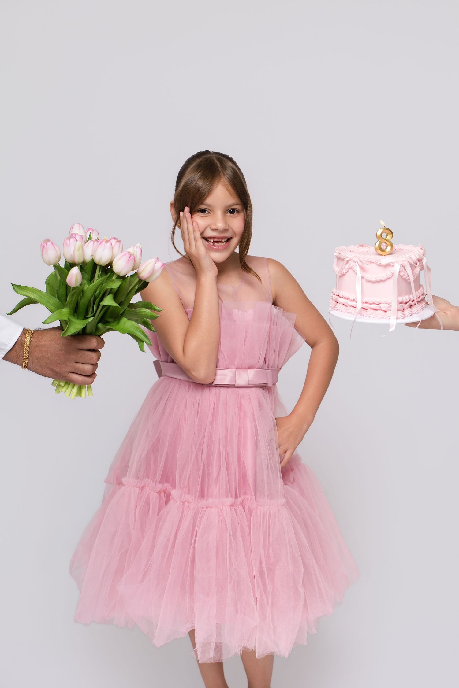 Young girl in pink tulle dress receiving tulip bouquet and birthday cake with number 8 candle on white background.