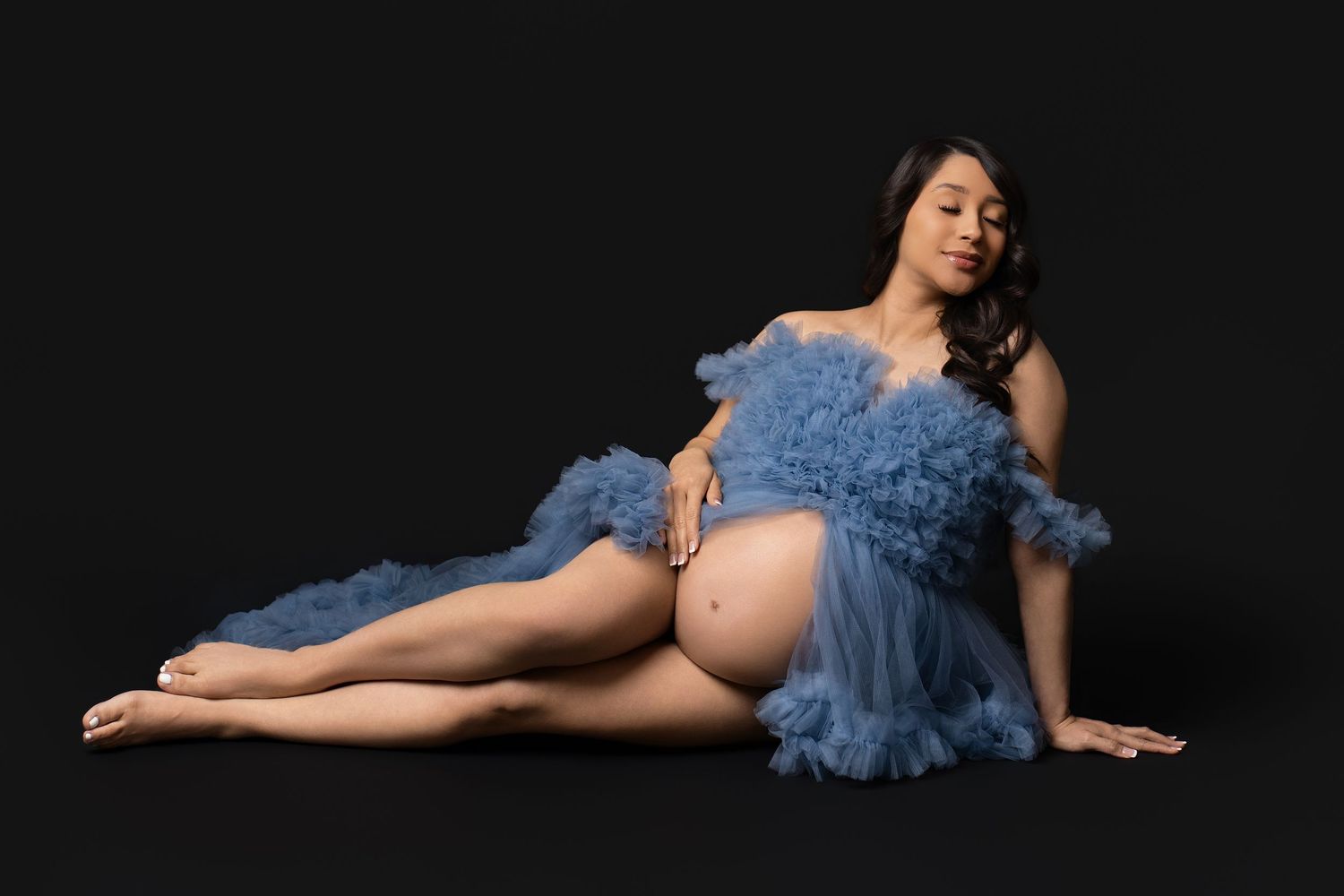 A woman in a blue tulle dress poses gracefully on a dark background in a boudoir photography style.