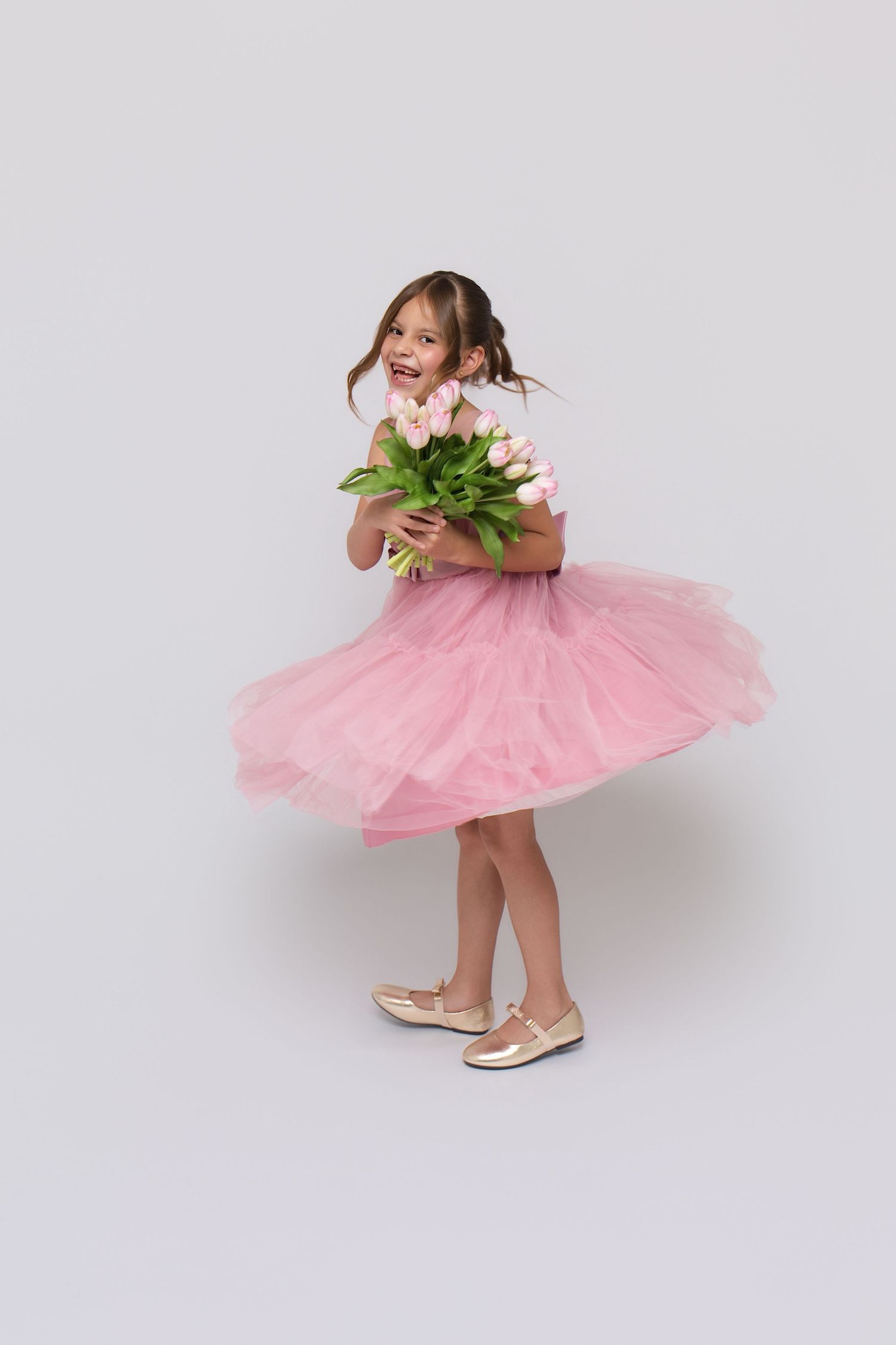 A joyful little girl in a pink tulle dress twirls while holding a bouquet of pink tulips on a white background.