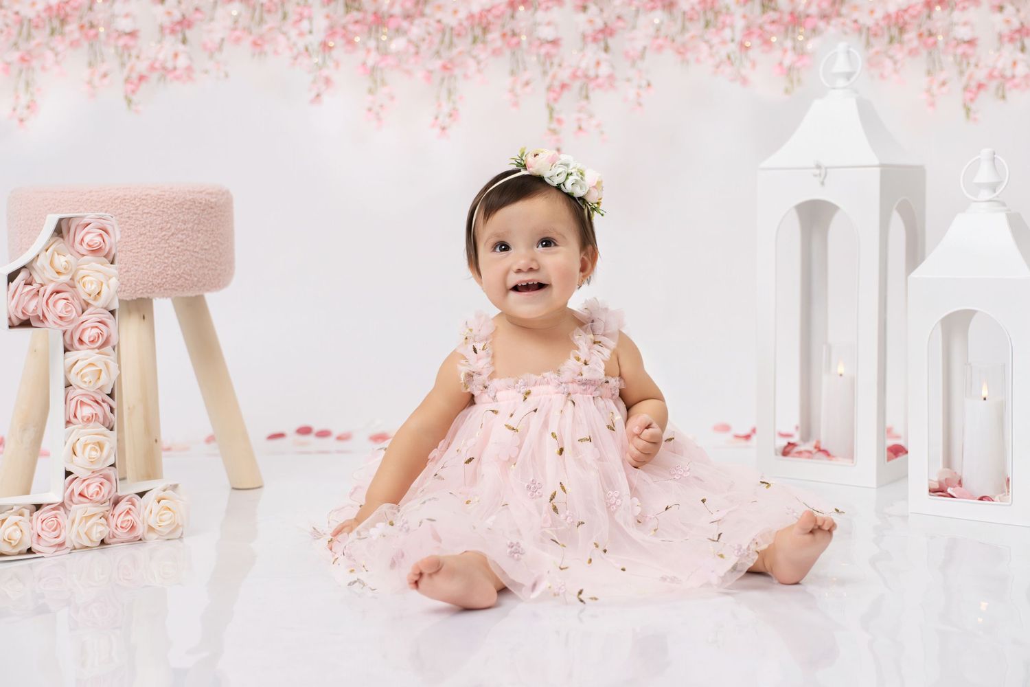 Baby girl in pink dress with floral crown sits beside number one decorations for a first birthday photo session.