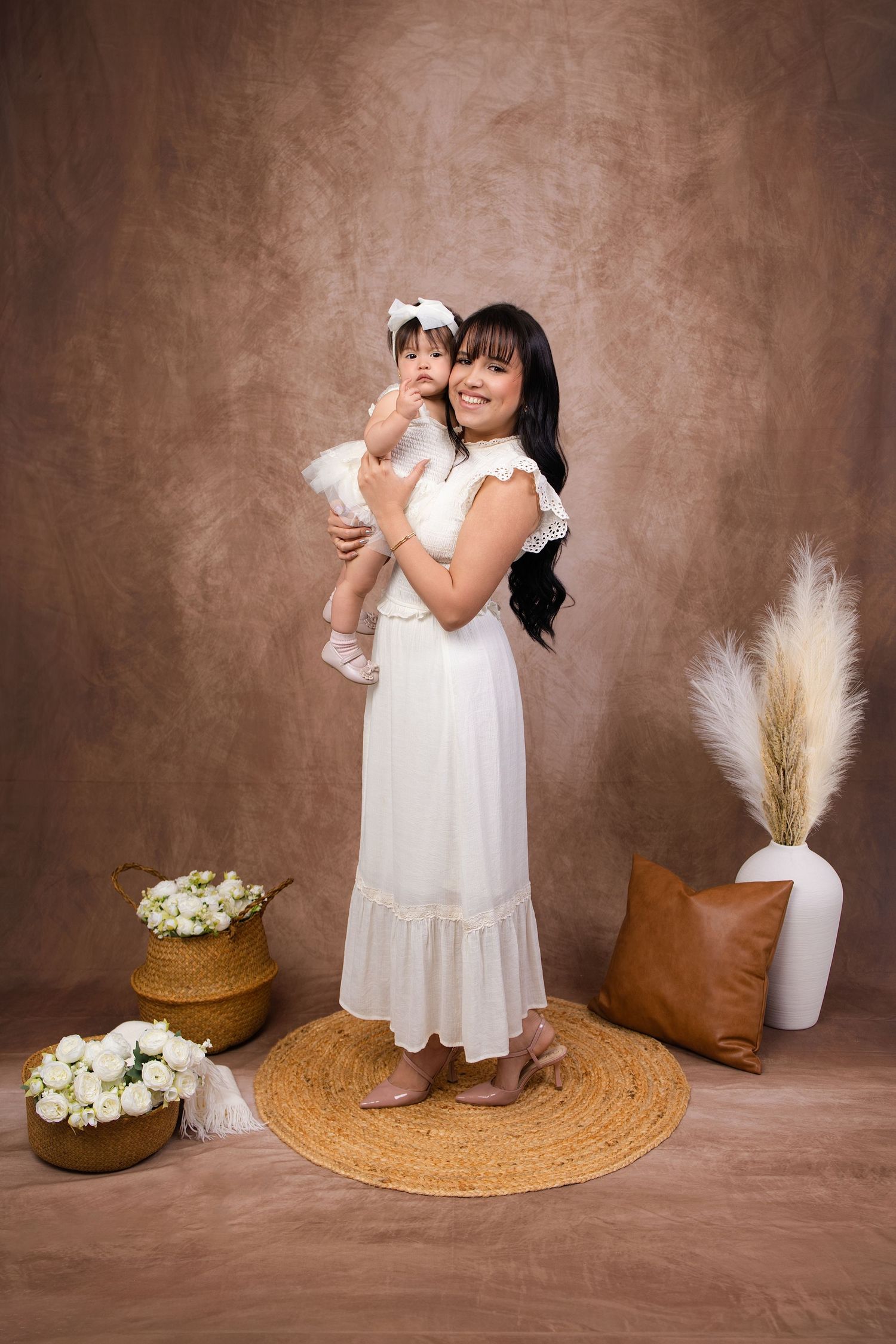 A smiling woman in a white dress holds a baby girl in a studio portrait with floral decor.