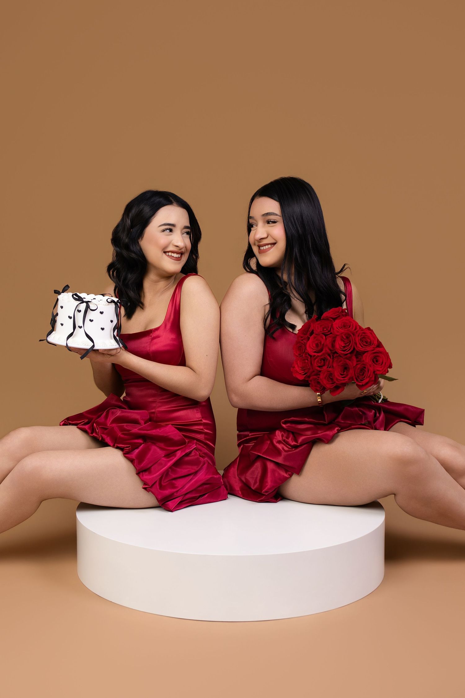 Two women in red satin dresses sit back-to-back holding a gift box and red roses bouquet on a beige background.