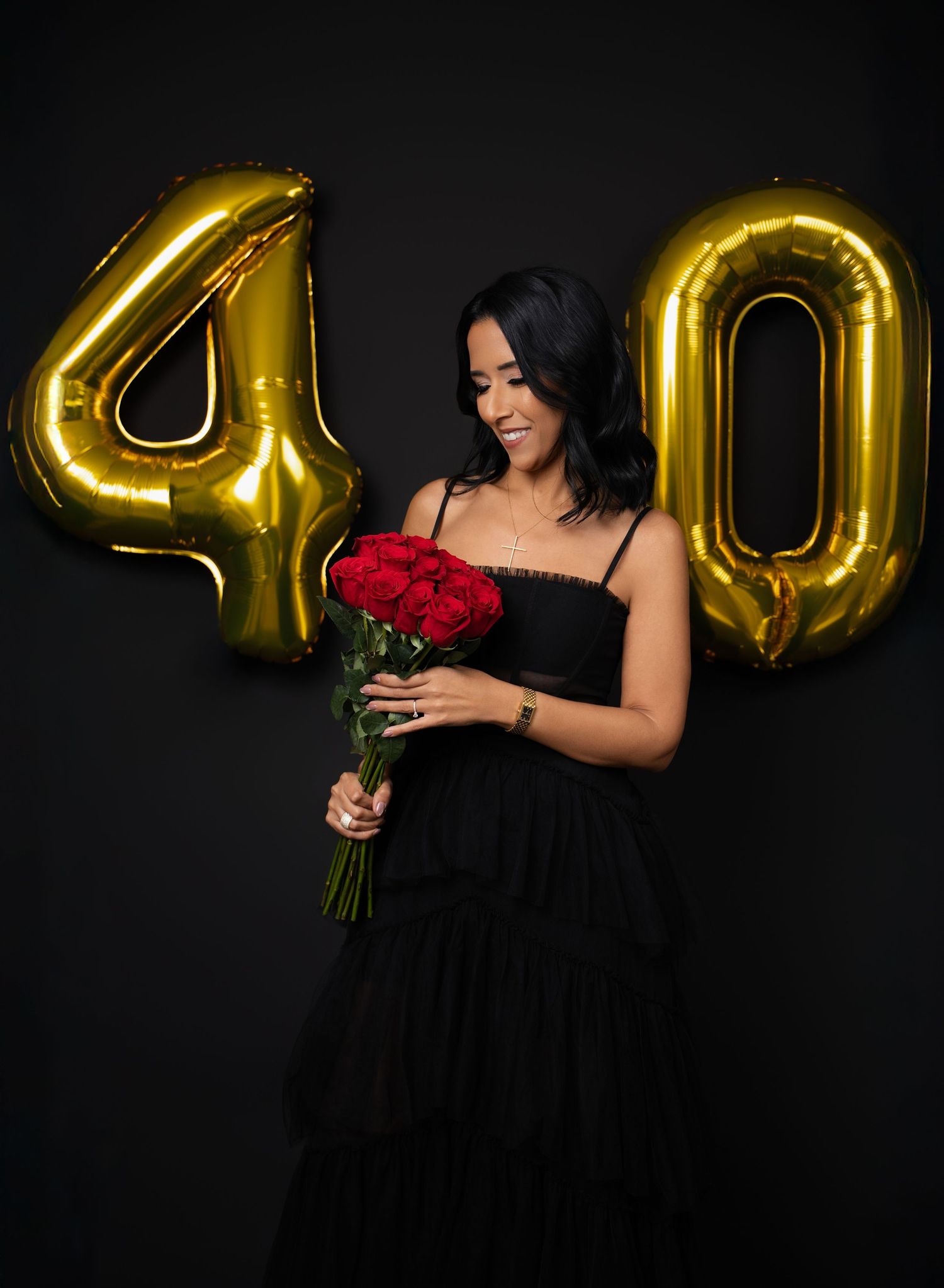 Woman in black dress holding red roses poses in front of gold number 40 balloons on dark background.
