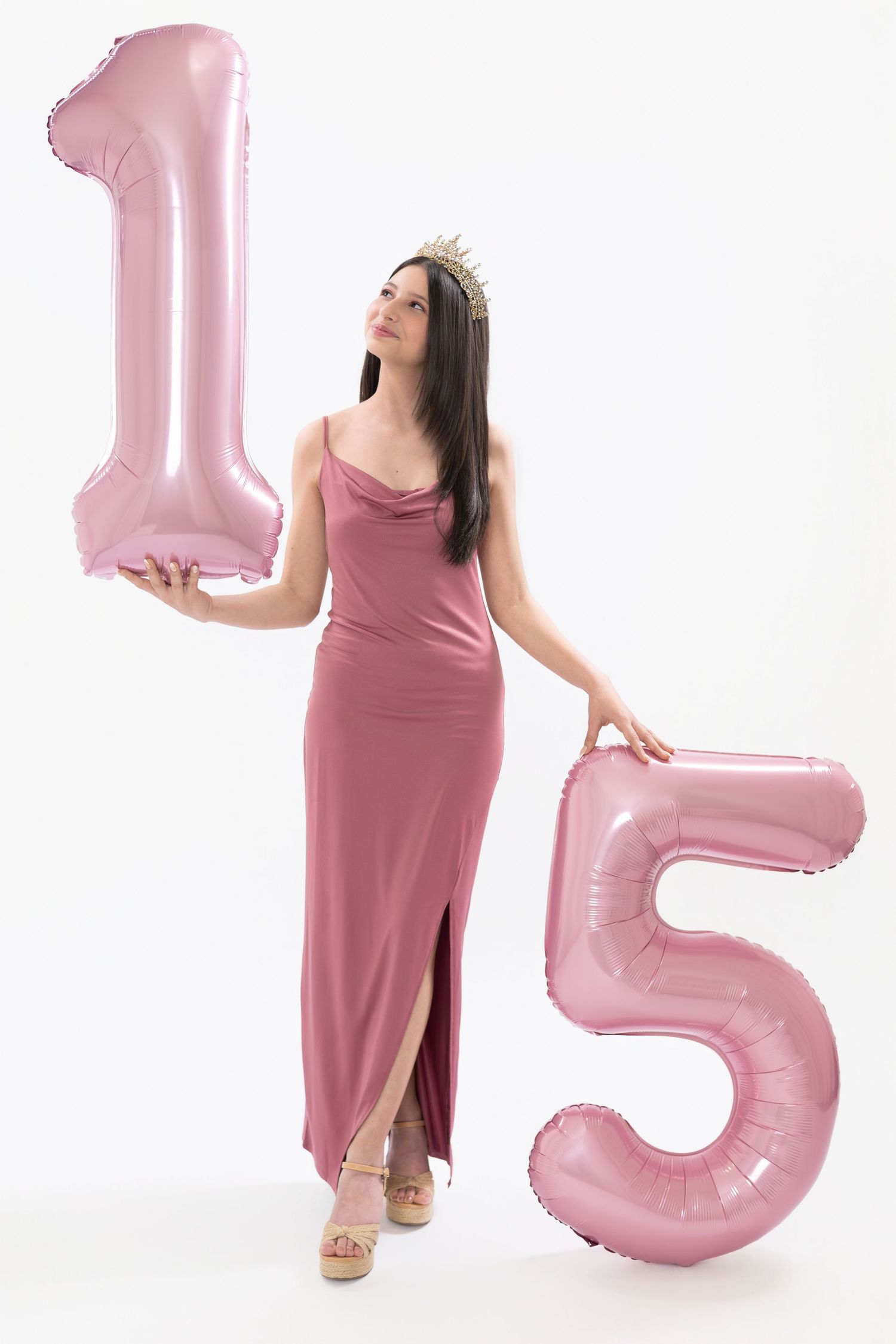 Young woman in pink gown and crown holds number 15 balloons, celebrating her quinceañera against white background.