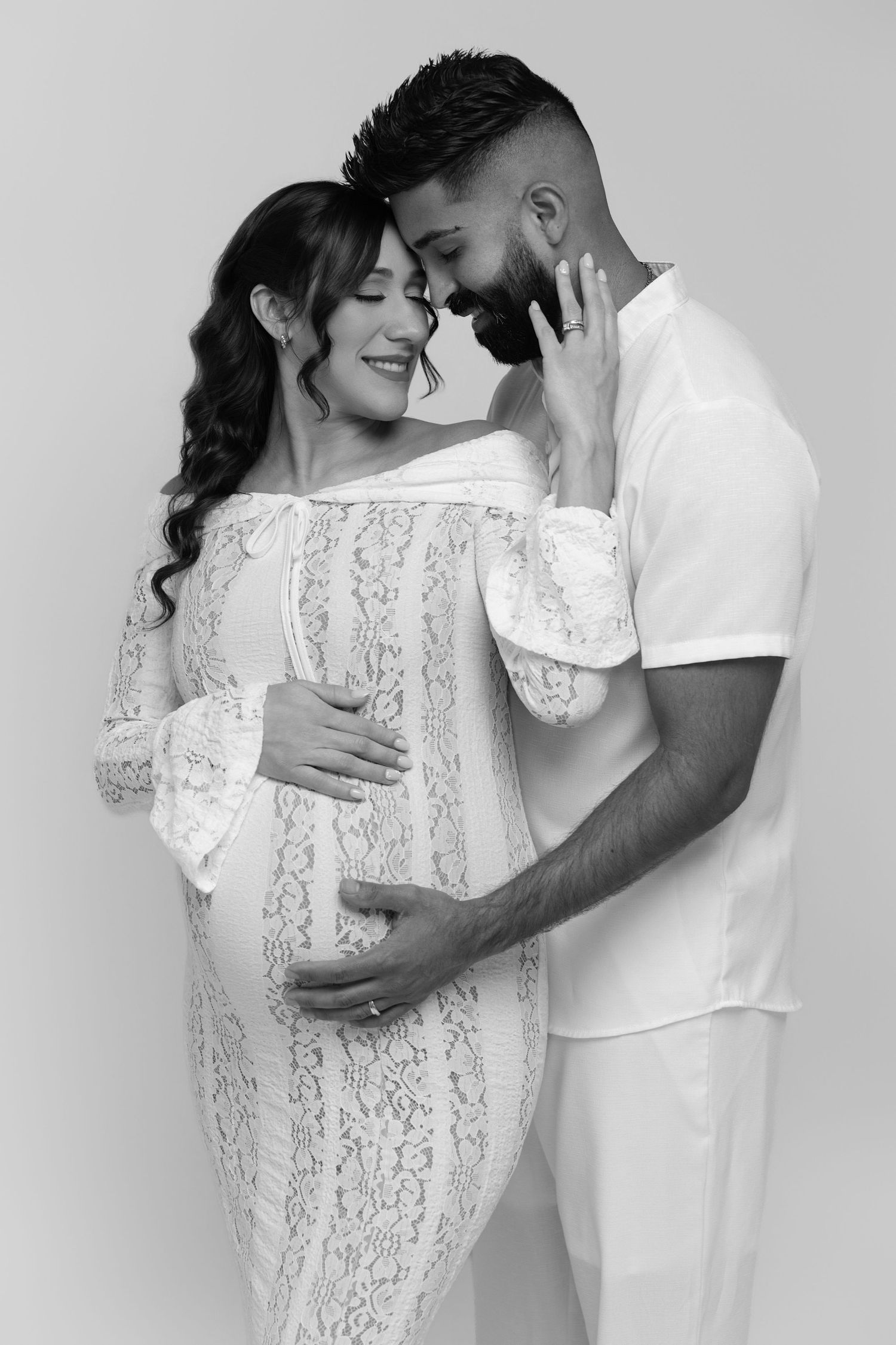 Black and white photo of a pregnant couple embracing tenderly, woman in lace dress, man in white outfit.