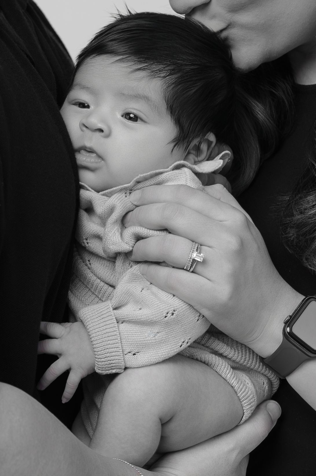 Black and white photo of a newborn baby being held and kissed by parents, wearing a knit sweater.