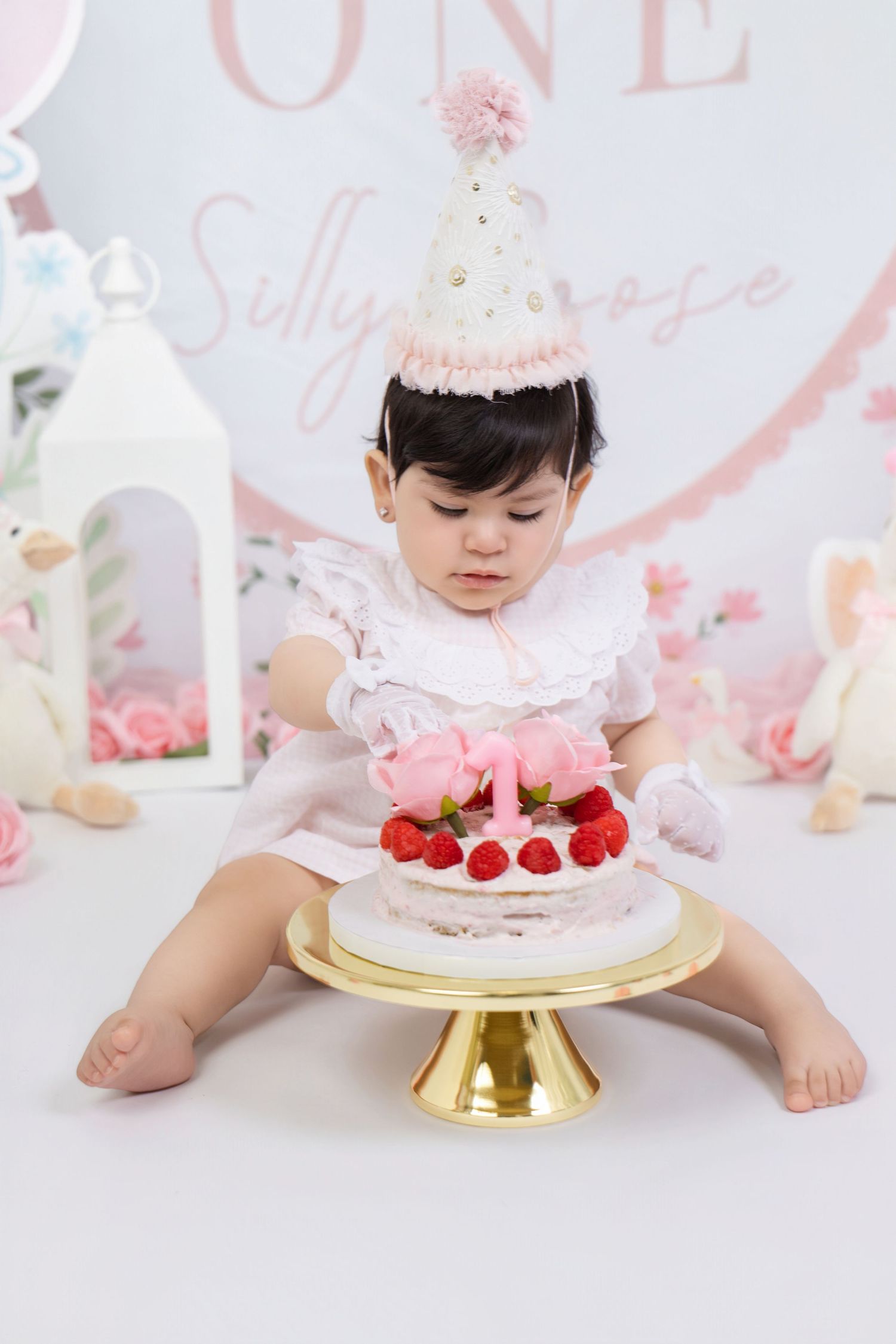 Baby girl in party hat reaching for pink cake on gold stand at first birthday cake smash photo session.