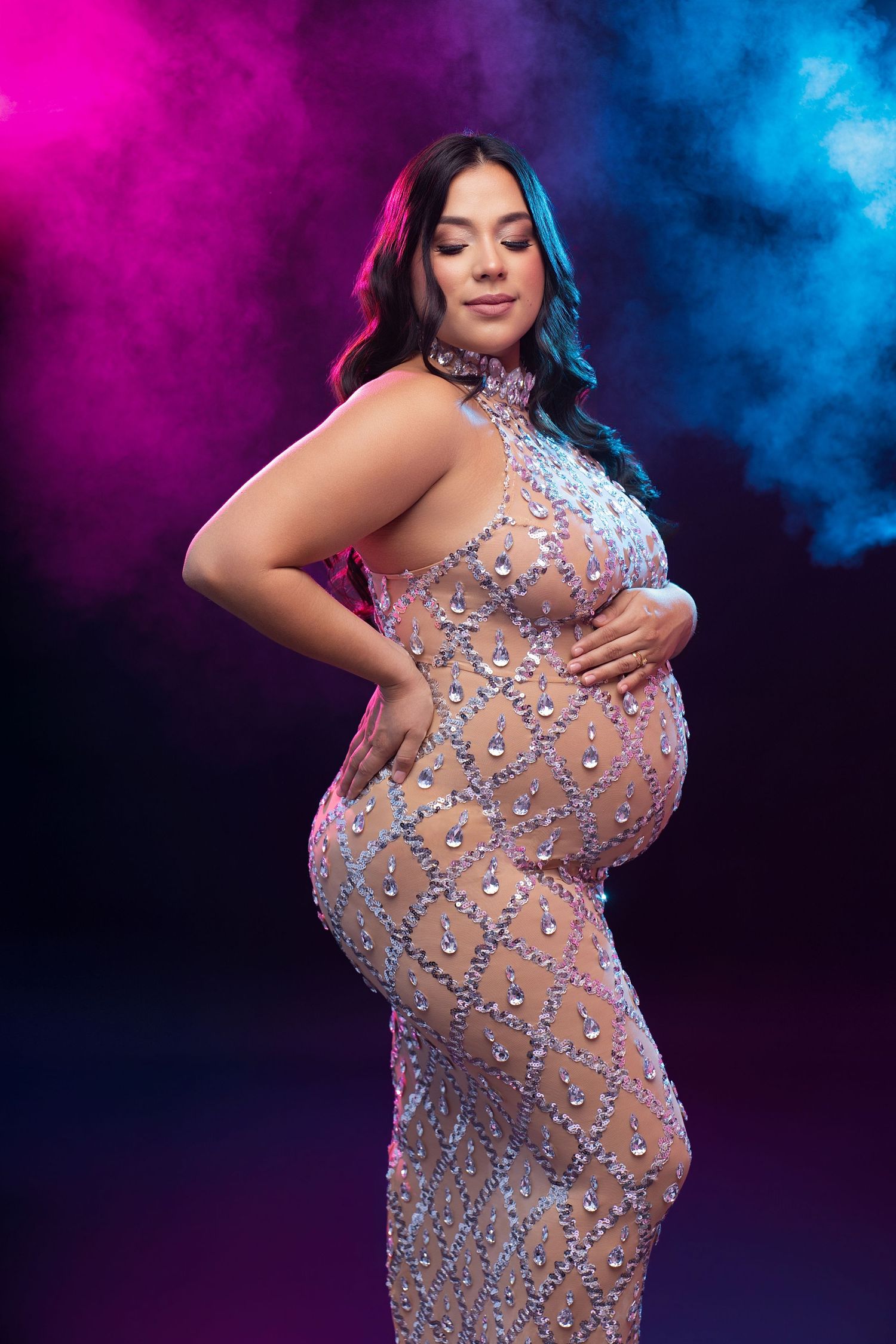 Pregnant woman in a sheer beaded gown poses against a dramatic pink and blue smoky background.
