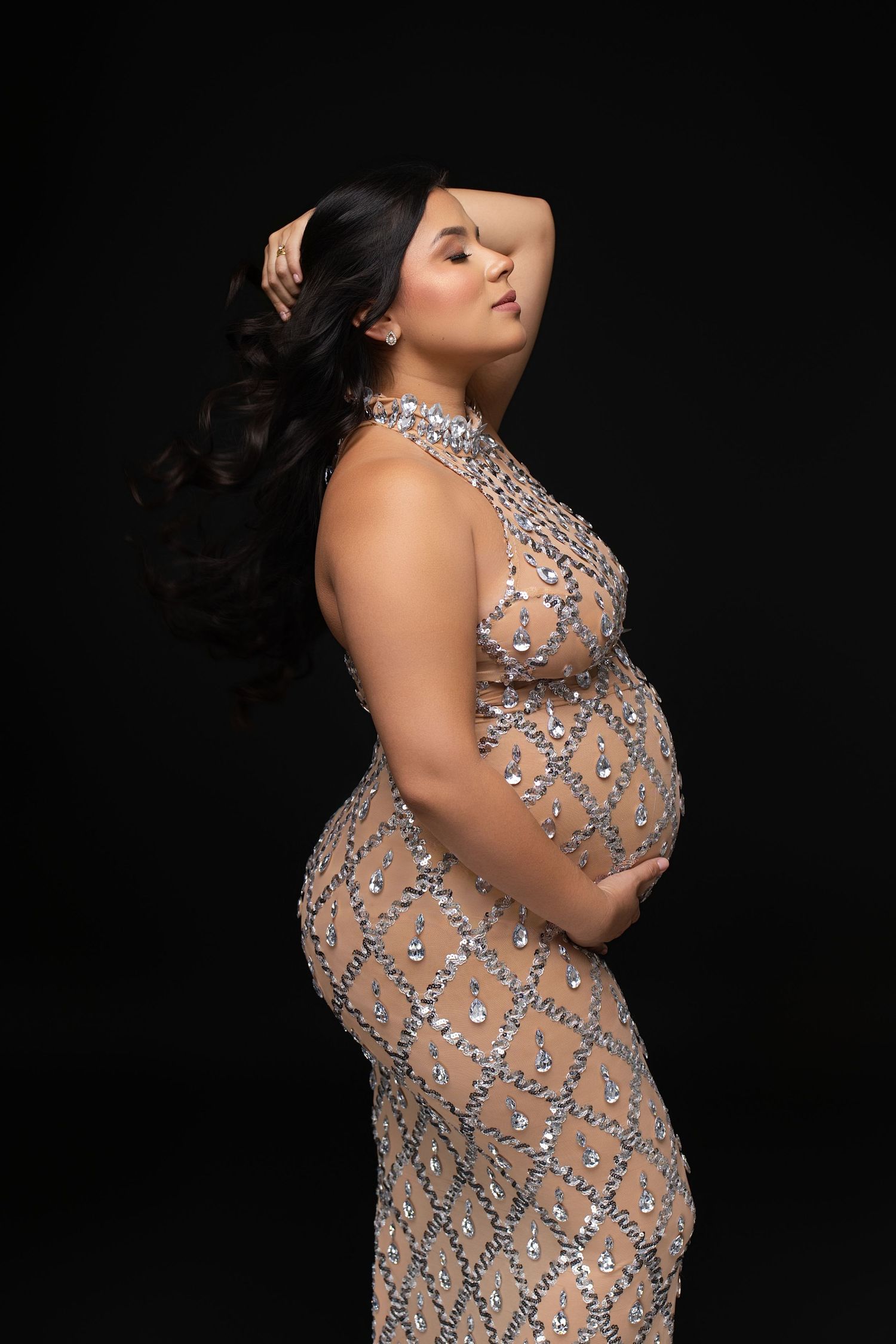 Pregnant woman in a beaded sequin gown poses elegantly against a black background, cradling her baby bump.