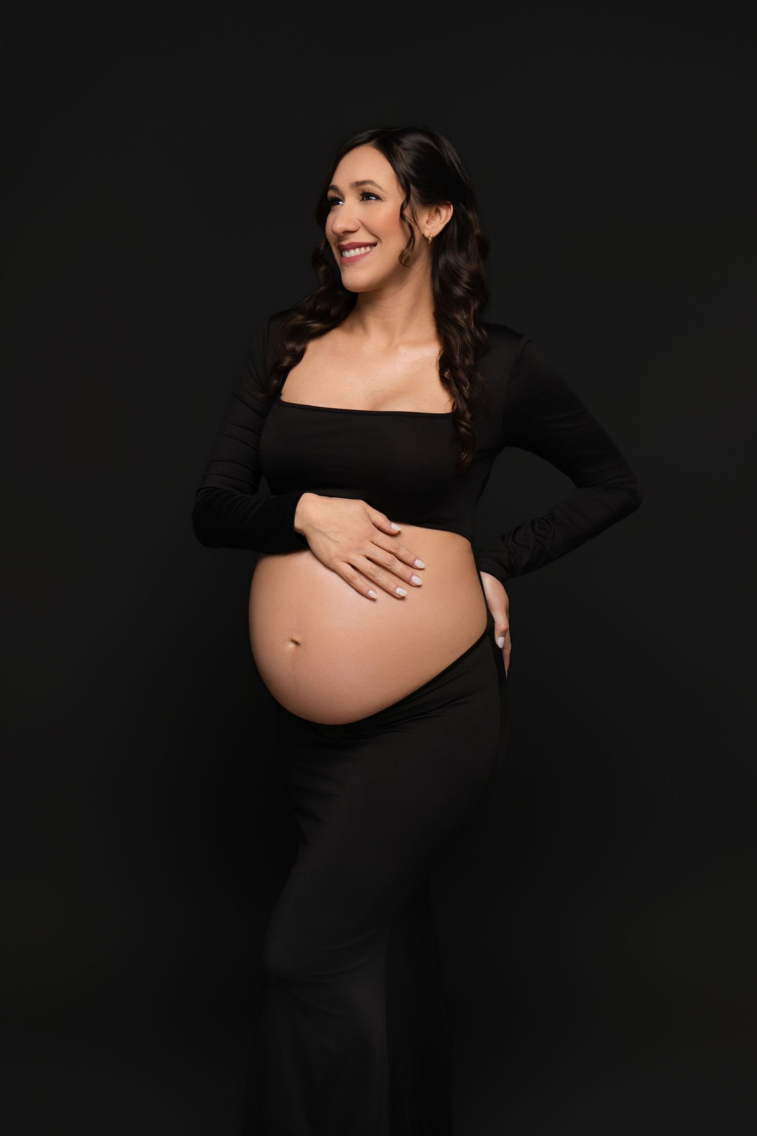 Smiling pregnant woman in black crop top and pants cradling her baby bump against a dark background.