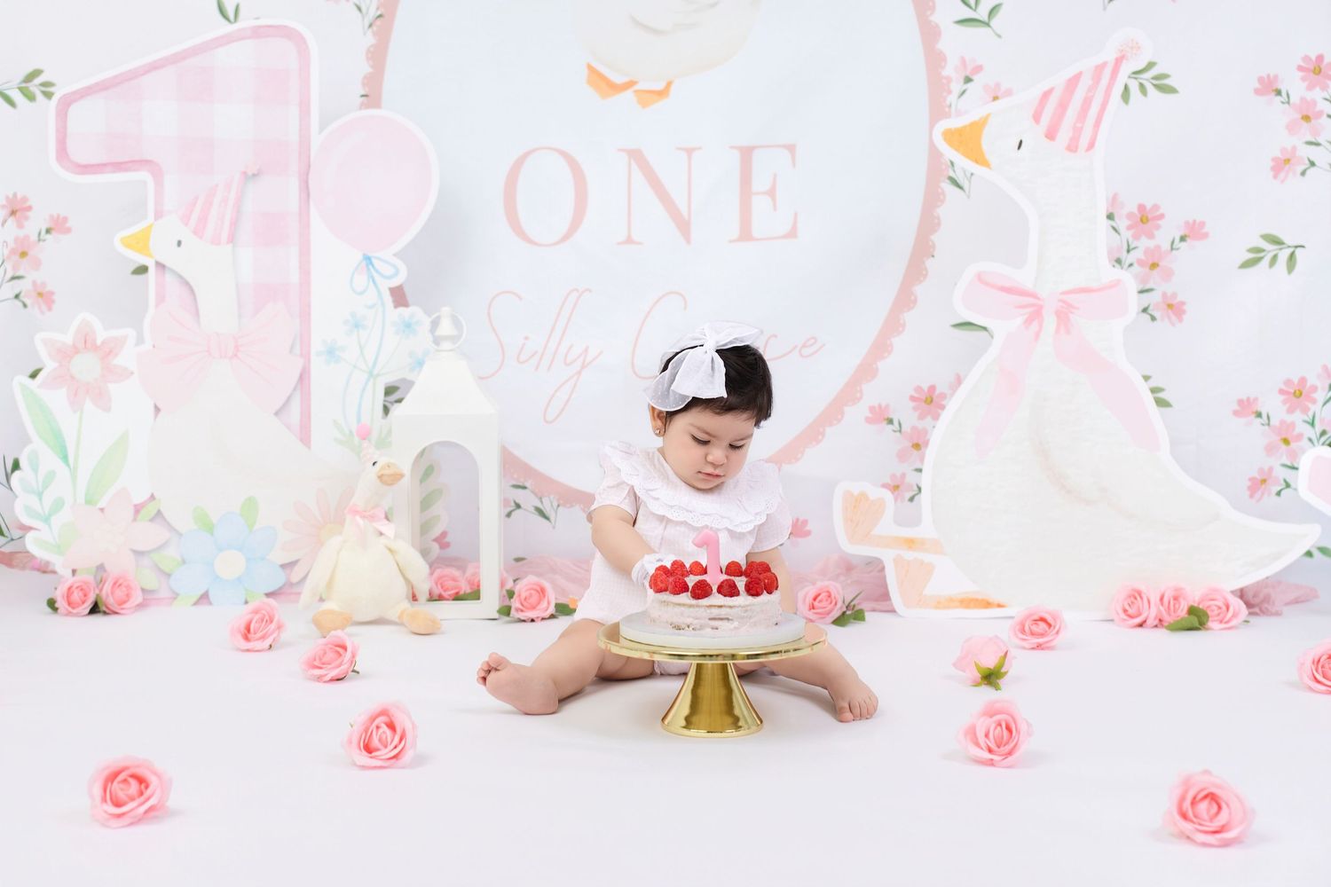 Baby girl in white outfit celebrates first birthday smash cake with strawberries, pink roses, and swan decorations.
