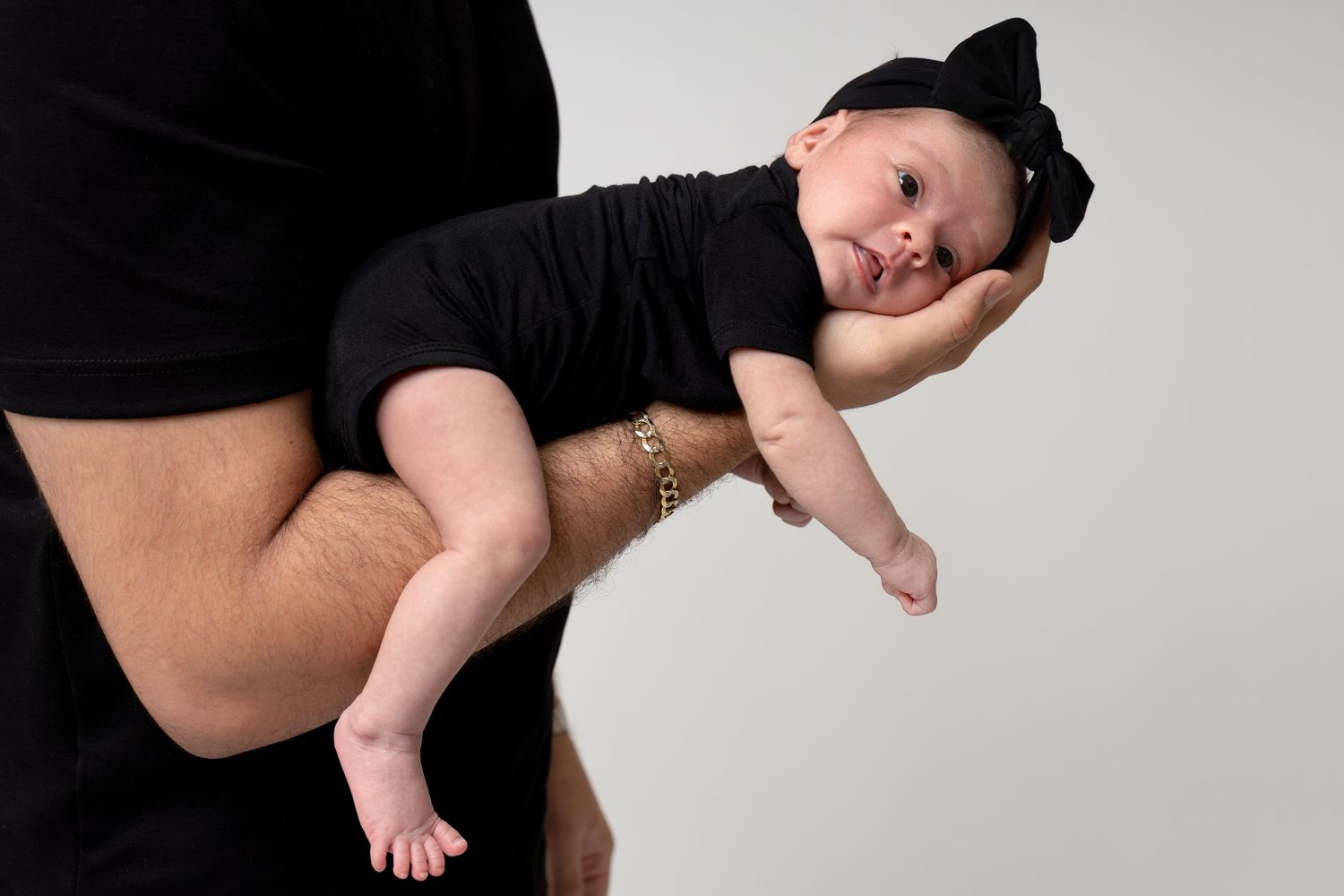 Adult holding a newborn baby dressed in black outfit with cat ear headband against a light gray background.