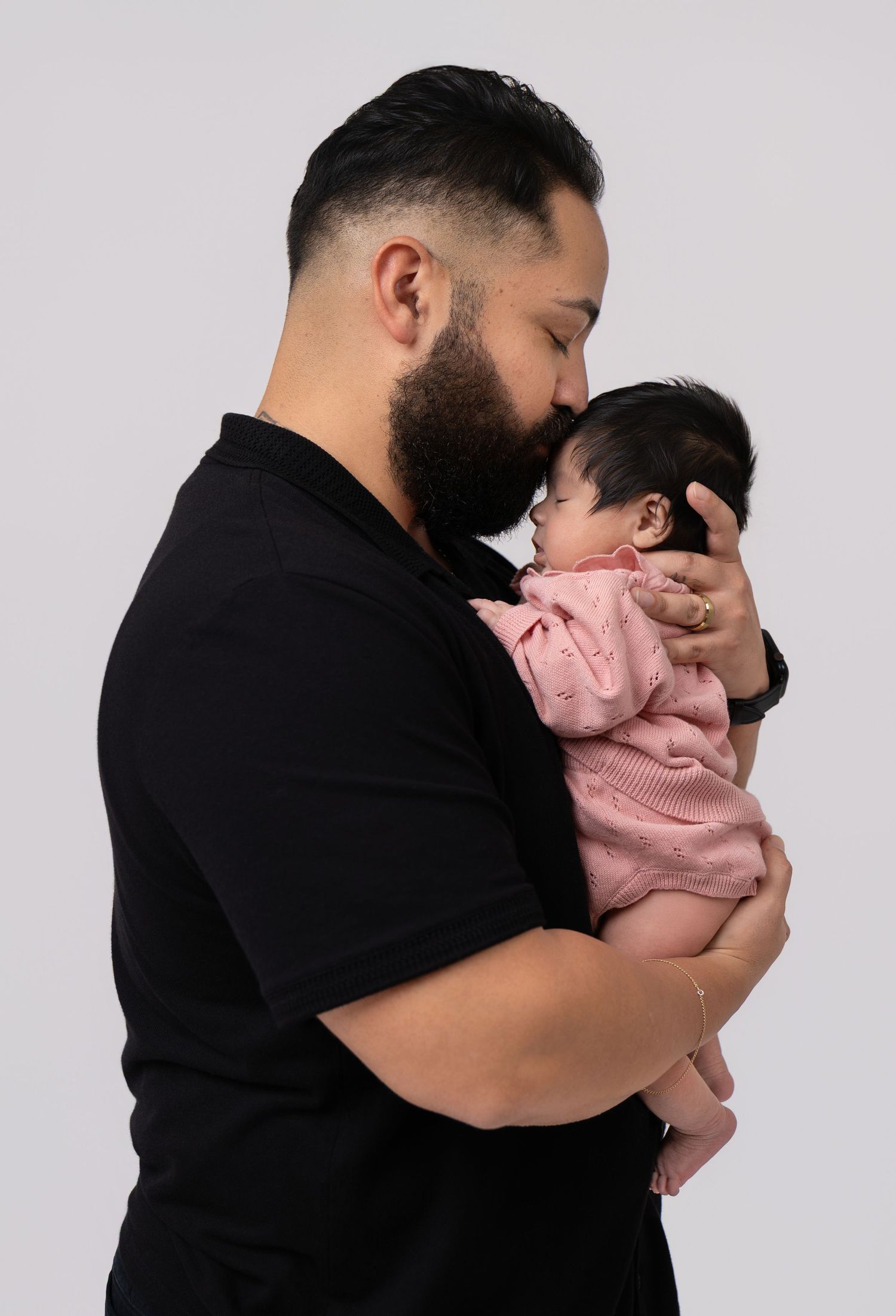 A bearded man in black shirt tenderly kisses a newborn baby dressed in pink, against a grey background.