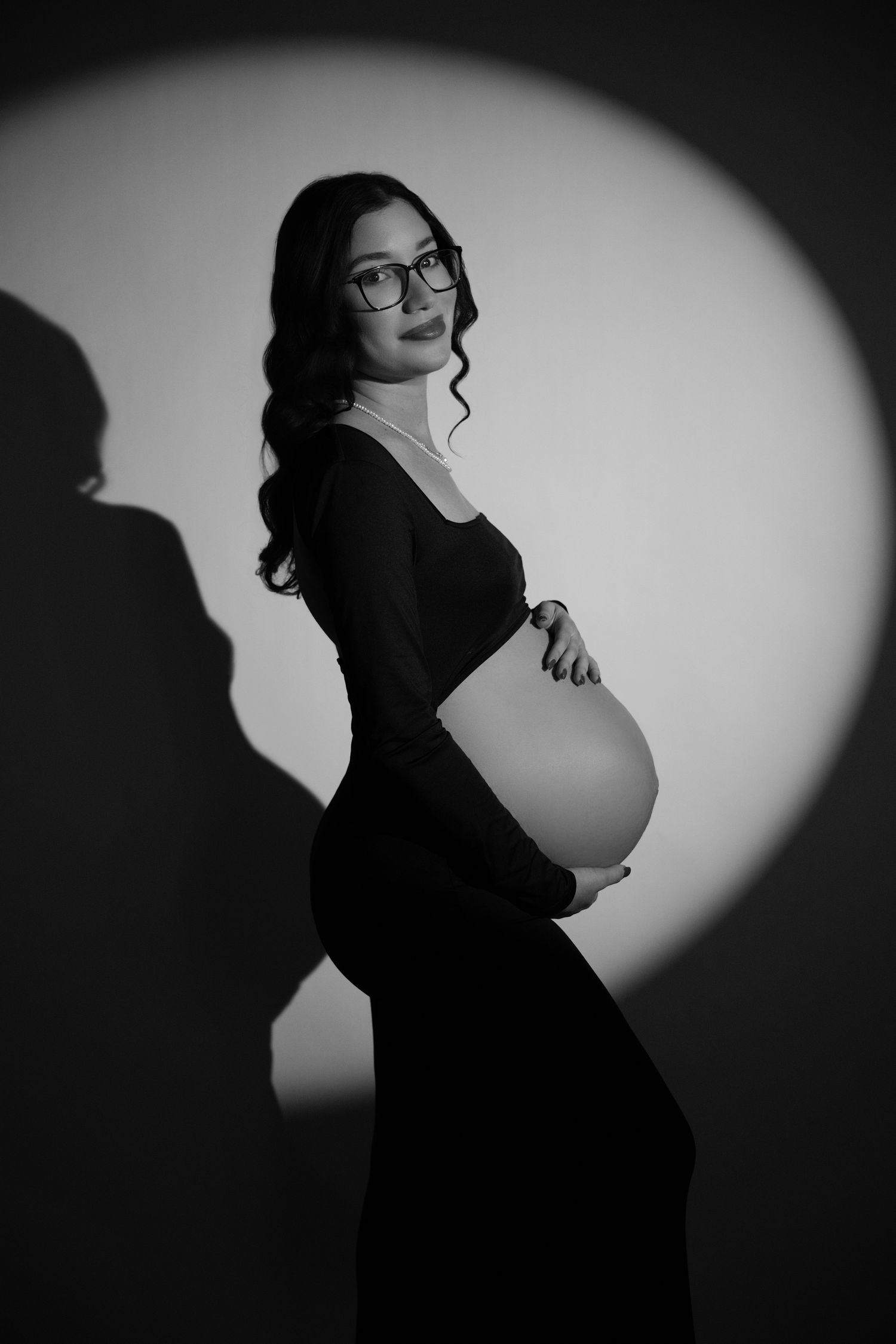 Black and white studio portrait of a pregnant woman in a black dress holding her baby bump under a circular spotlight.