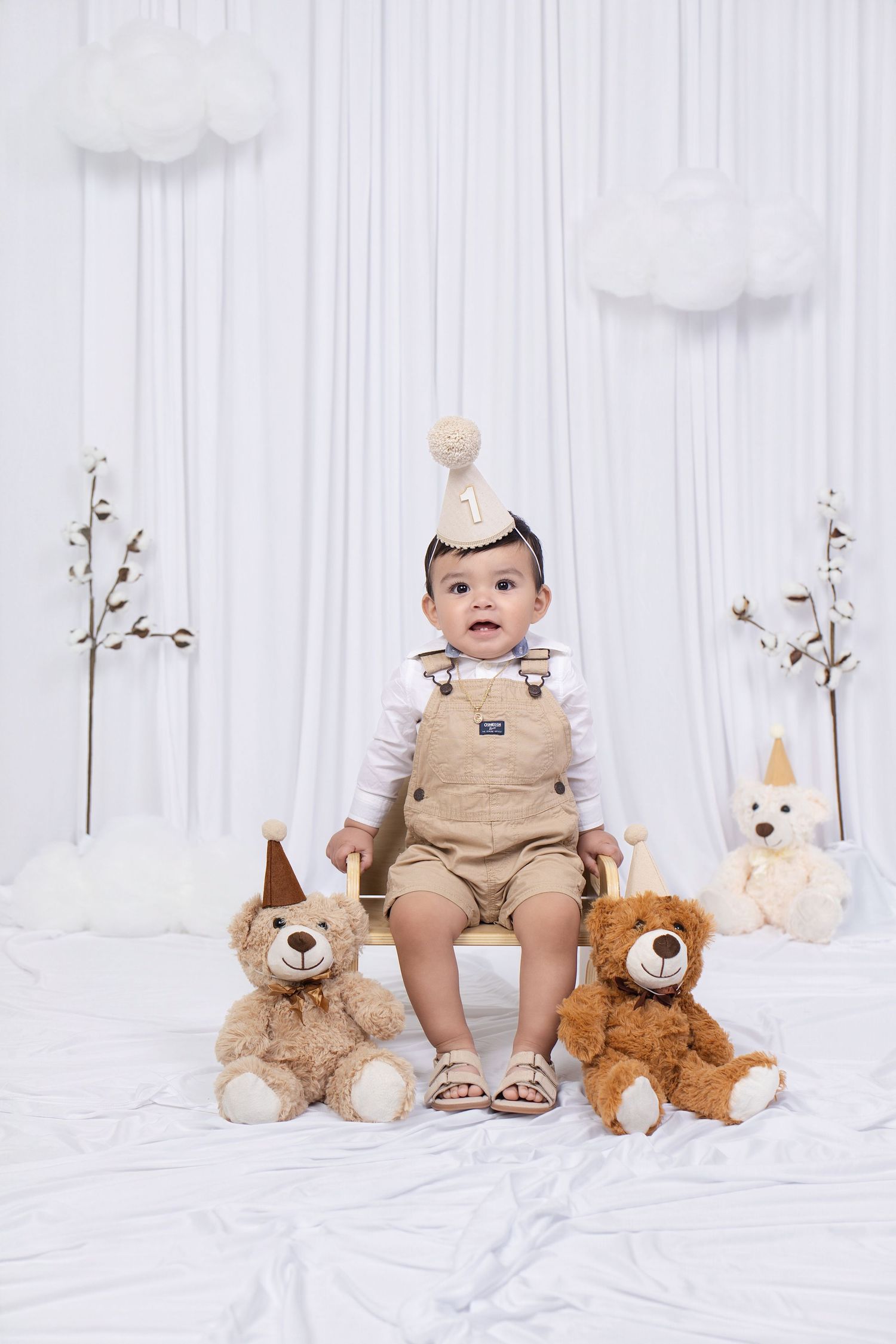 Baby boy in tan overalls and party hat sits on a box surrounded by teddy bears at a first birthday photo shoot.