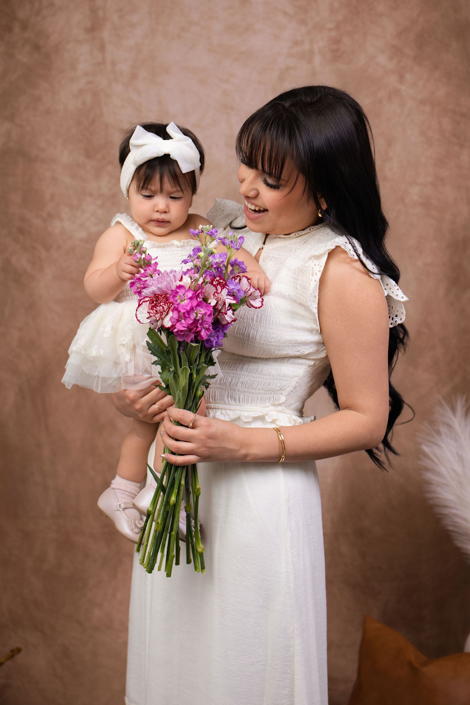 A woman in a white dress holds a baby girl and a bouquet of pink and purple flowers in a studio portrait.