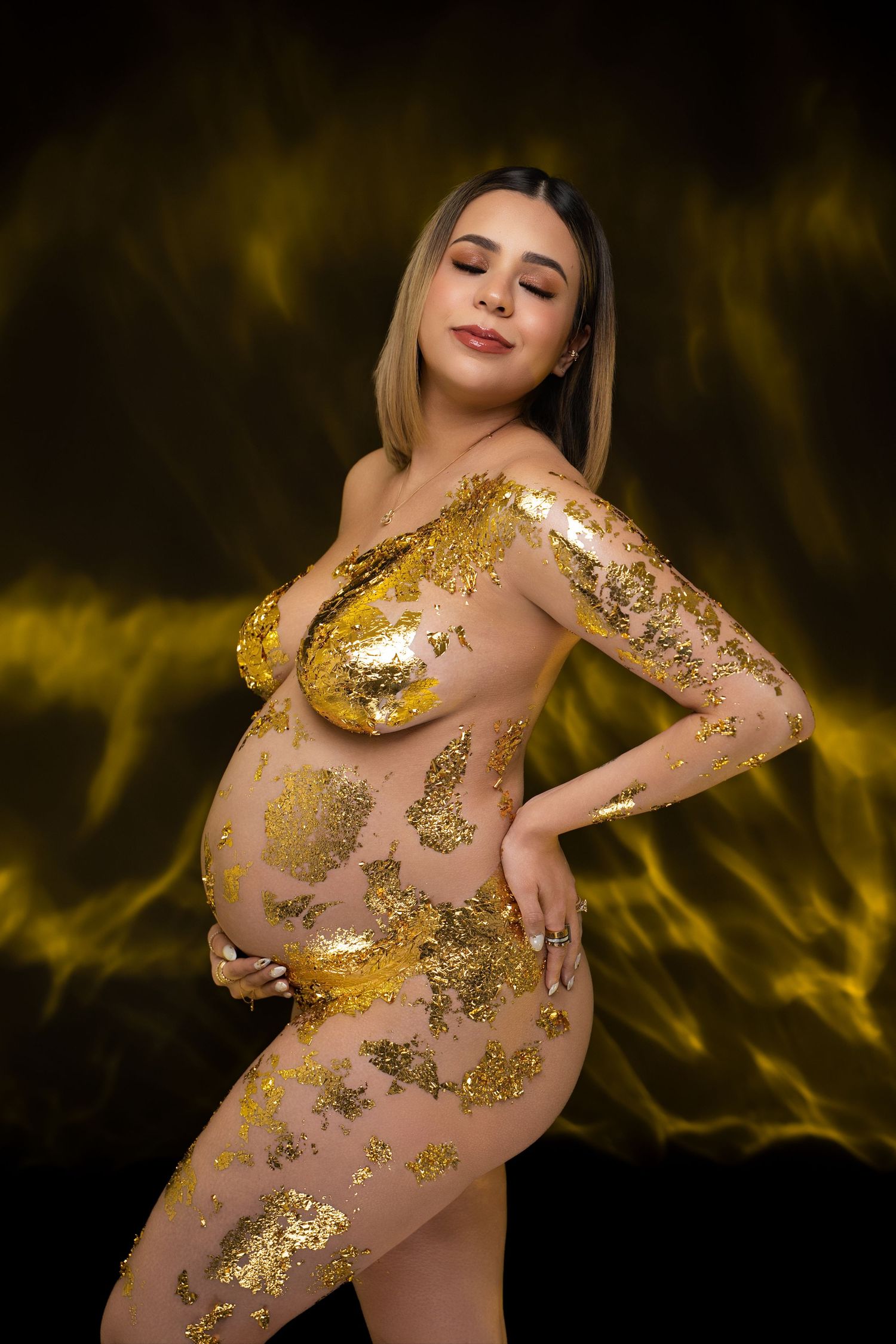 Pregnant woman posing with gold foil body art against a dramatic black and yellow background.