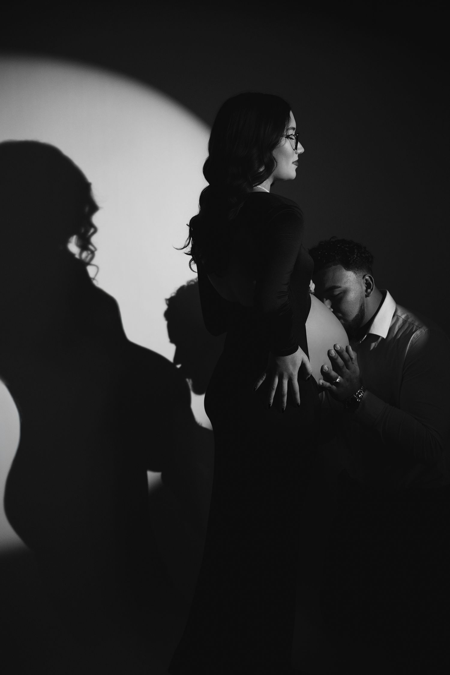 Black and white maternity photo of a pregnant woman with her partner kneeling beside her, dramatic shadows on wall.