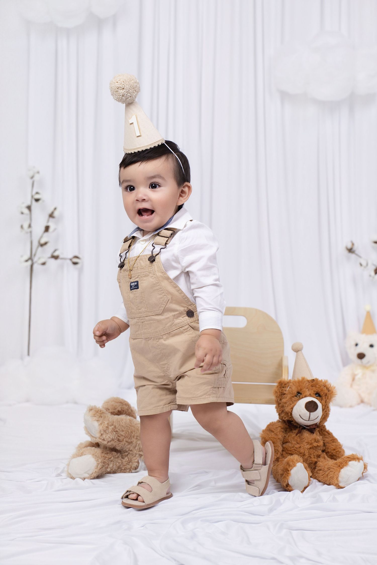 A baby boy in beige overalls and a birthday hat stands between two teddy bears in a white studio setting.