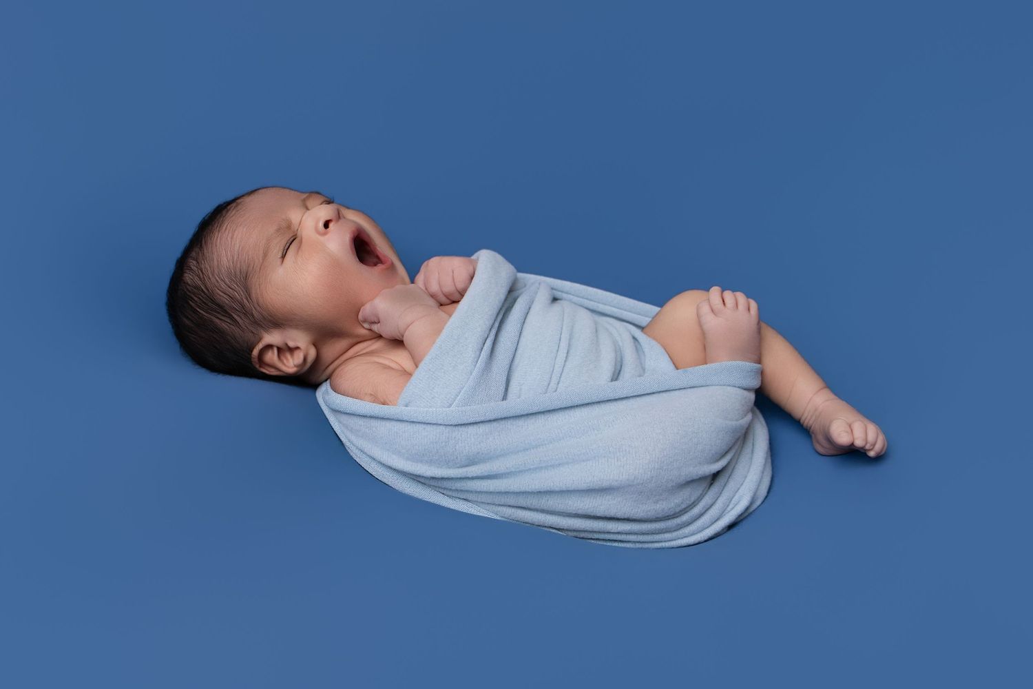 Newborn baby swaddled in light blue wrap, yawning on a blue background.