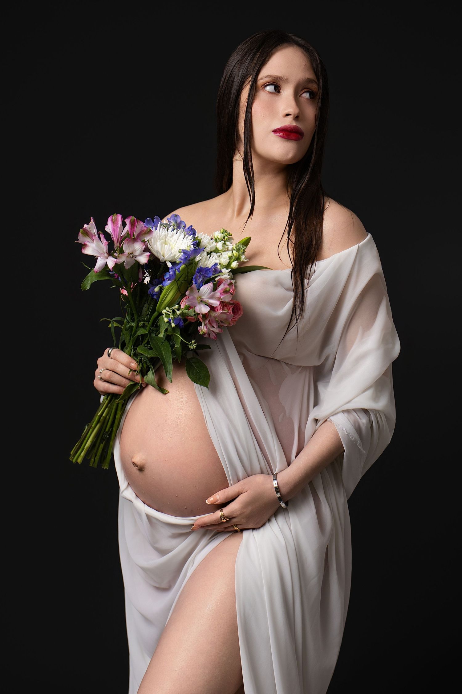 Pregnant woman in sheer white wrap holding colorful flower bouquet against dark background.
