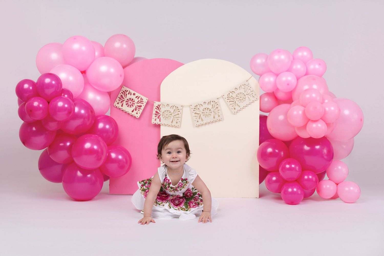 Baby girl sits smiling among pink and magenta balloon garlands with a cream arch backdrop at a first birthday celebration.