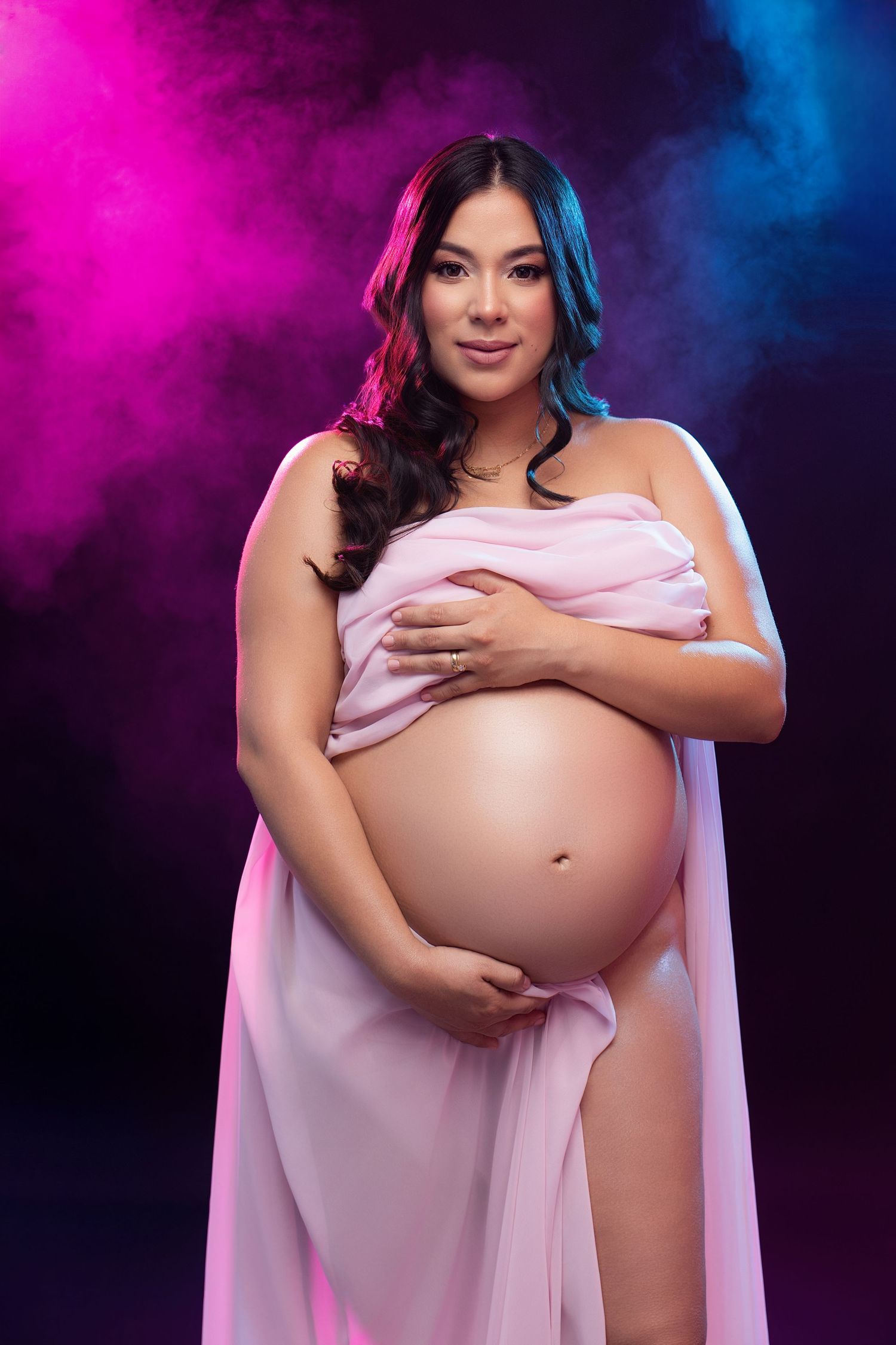 Pregnant woman in pink draped fabric cradling baby bump against dramatic pink and blue smoky background.