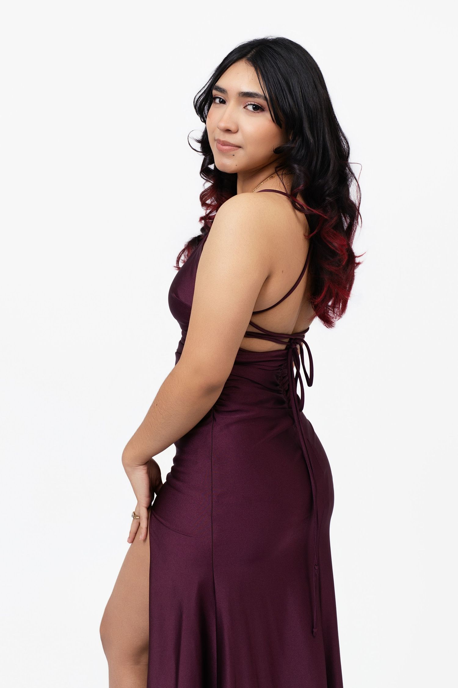 Woman in elegant burgundy backless satin gown with thigh slit, posing gracefully against white background.