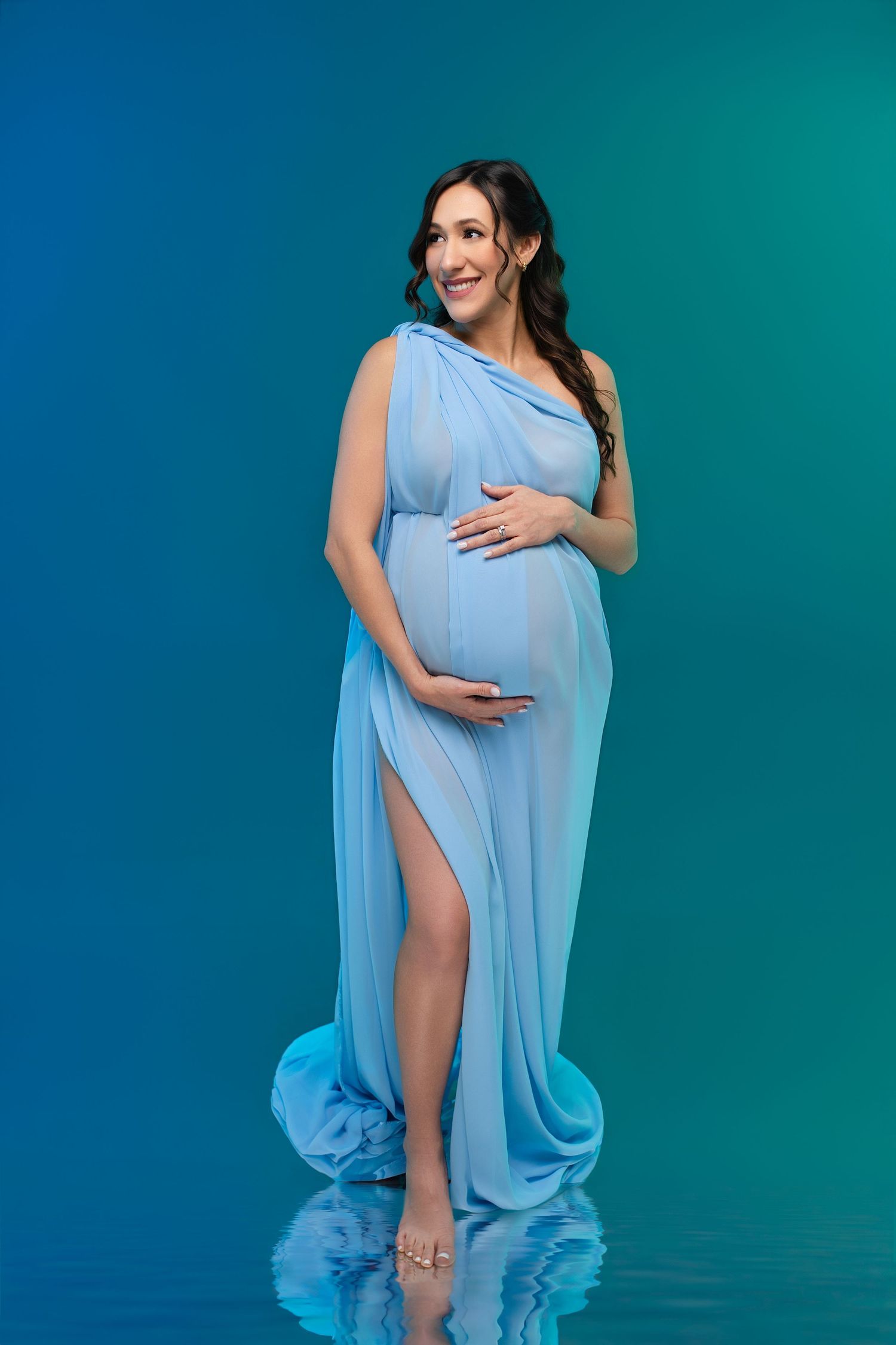 Smiling pregnant woman in elegant blue draped gown holding baby bump, standing on reflective surface.