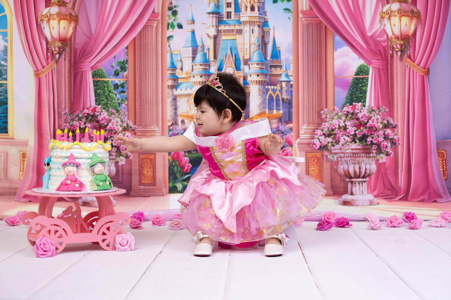 Toddler girl in pink princess dress reaches for birthday cake at Disney castle-themed first birthday photo shoot.
