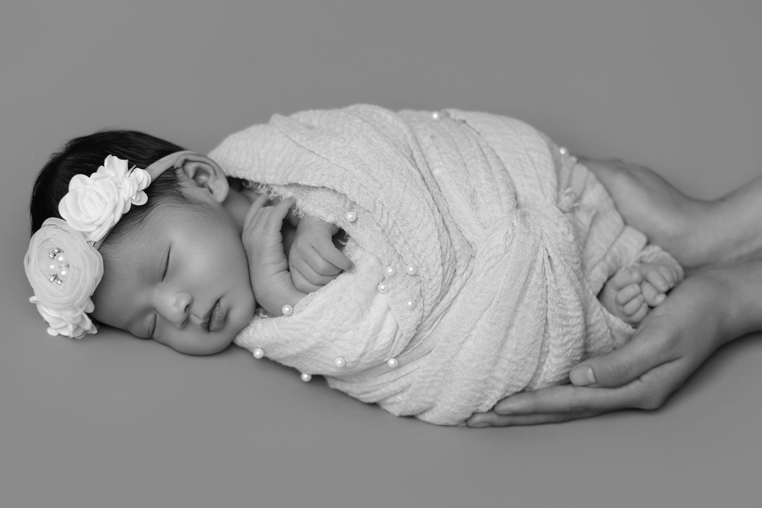 Black and white photo of a sleeping newborn baby girl swaddled in white wrap with floral headband in hands.