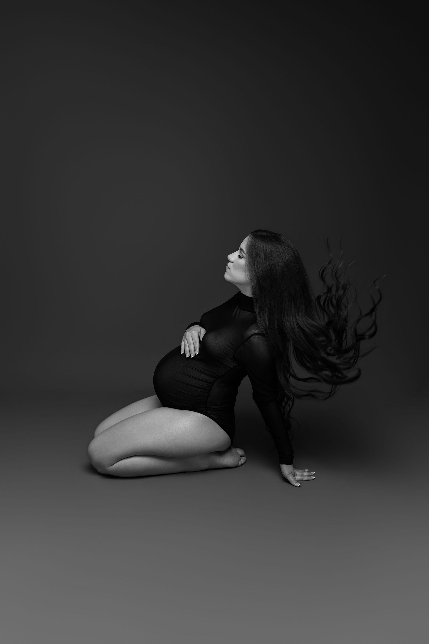 Black and white photo of a pregnant woman sitting gracefully with flowing hair, wearing a black outfit against a dark background.