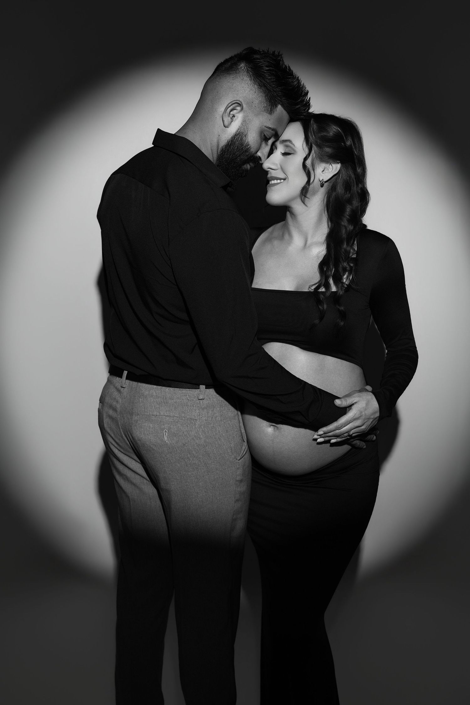 Black and white maternity photo of a couple embracing, man in black shirt holding pregnant woman's belly.