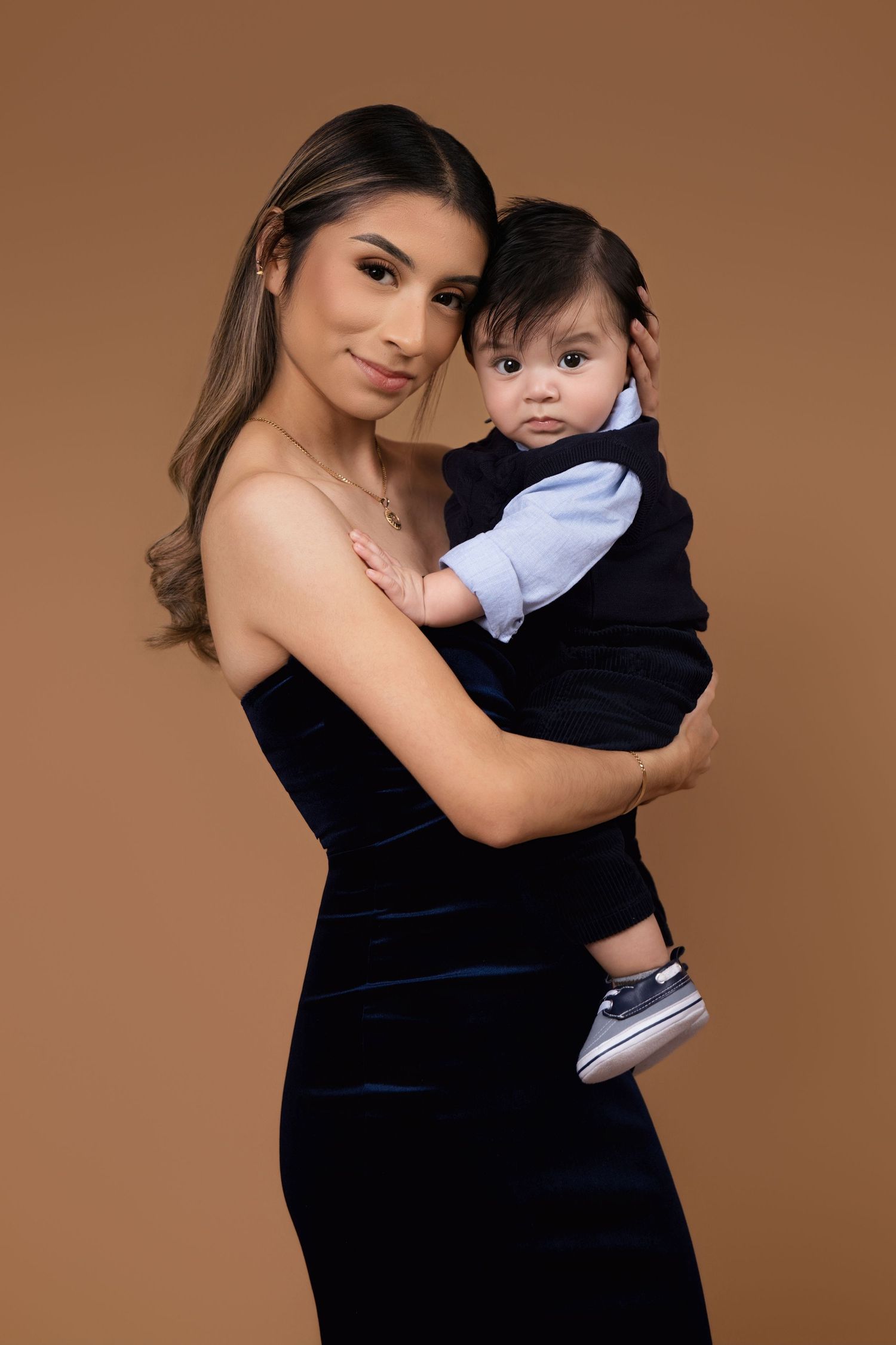 A parent and baby pose together against a brown backdrop wearing formal attire including a black dress and navy suit.