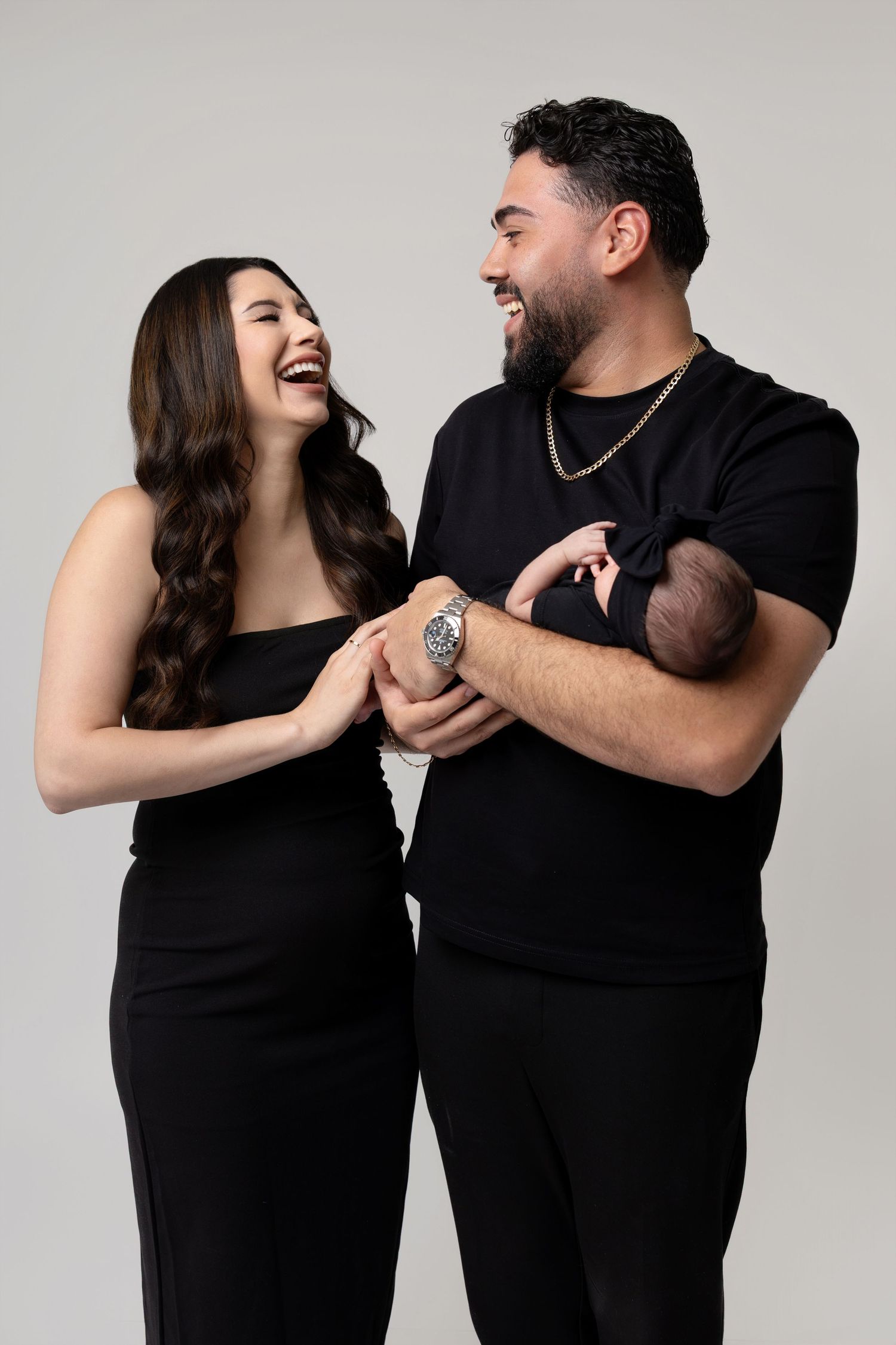 A joyful couple dressed in black holding hands and a newborn baby, laughing together against a gray background.