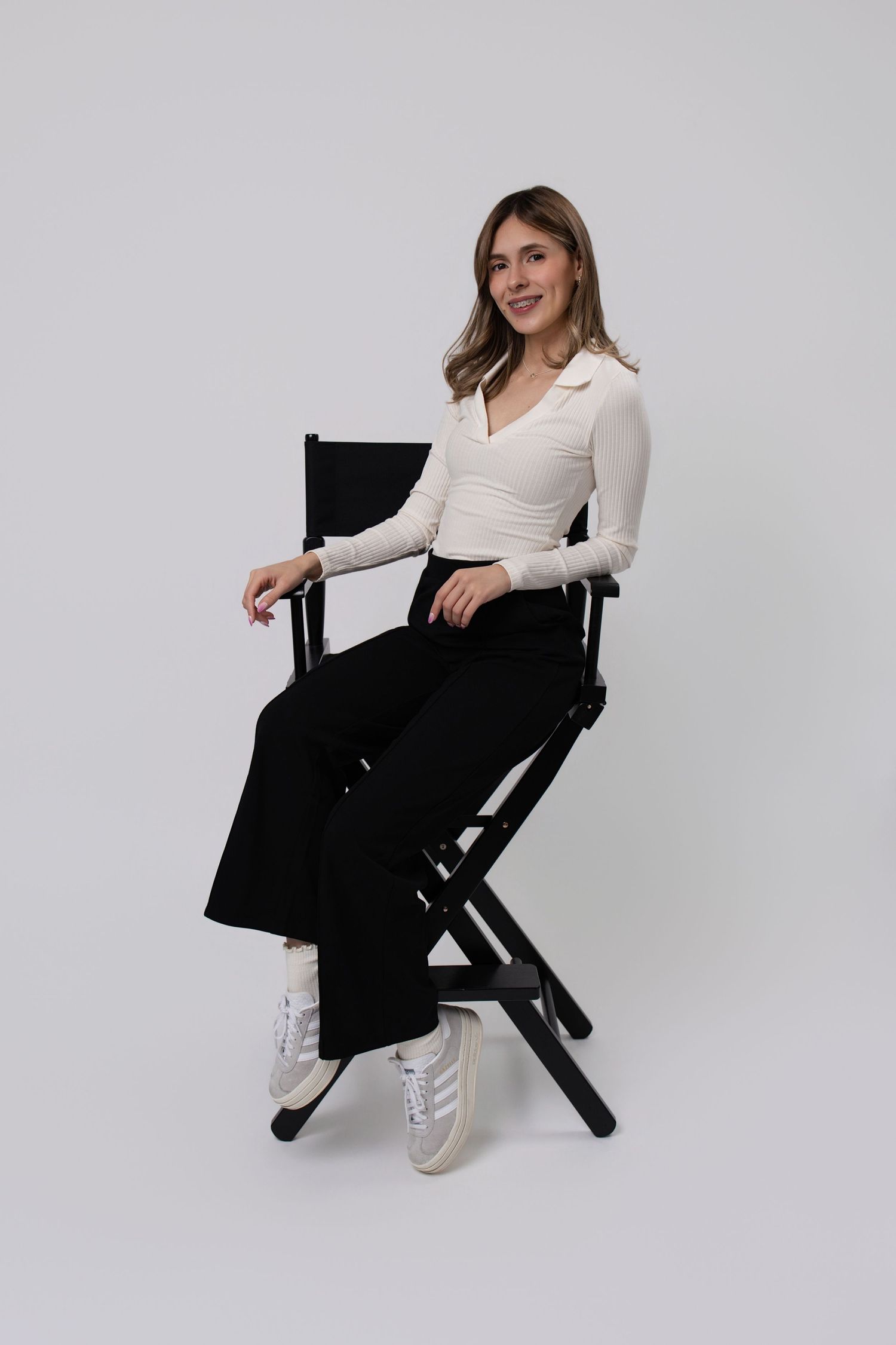 Woman in white top and black wide-leg pants sits on a director's chair, smiling against a gray background.